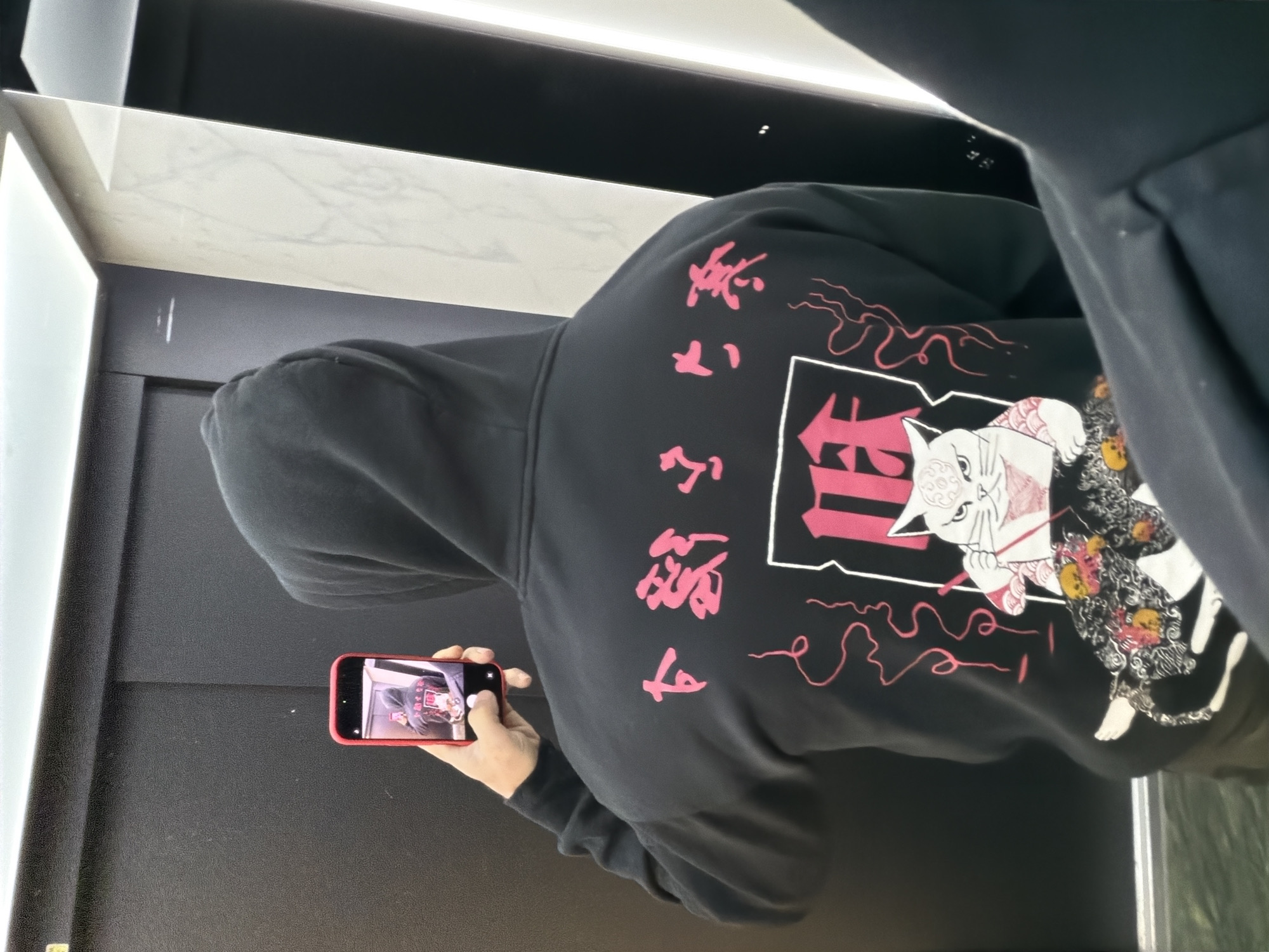 A person wearing a black hoodie with pink Japanese lettering and a cat illustration on the back is standing indoors, holding a red smartphone and taking a picture of themselves in a mirror. The location appears to be a store or dressing room with black walls and a white marble countertop. The city of Plovdiv, Bulgaria is not identifiable from the image.