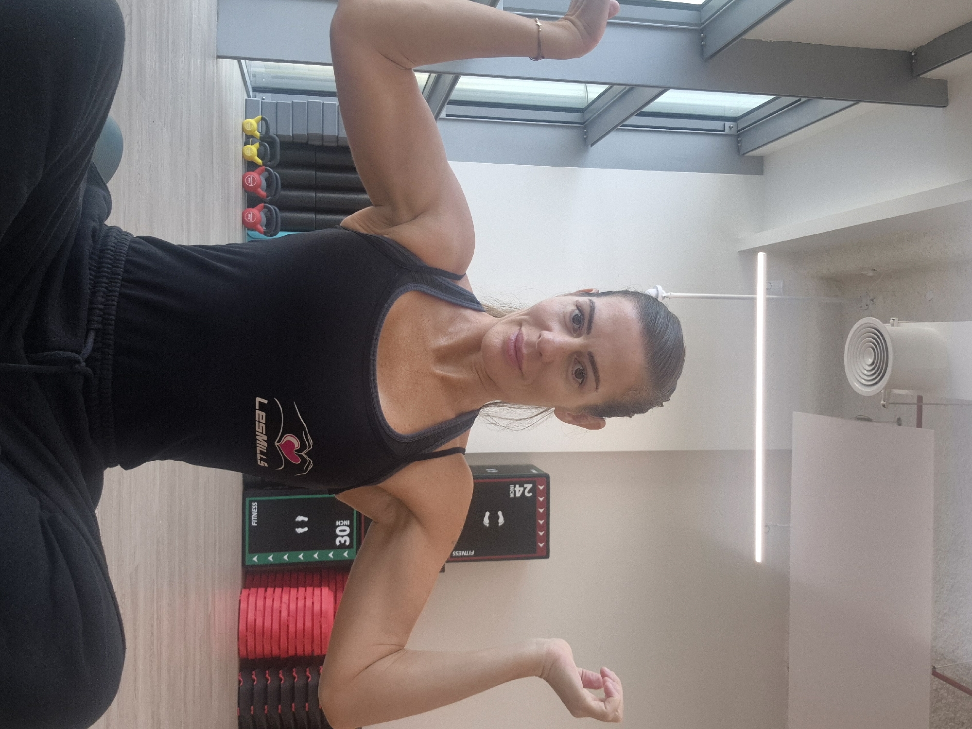 The image shows a woman in a gym, with a rack of weights in the background. She is wearing a black tank top and pants, and her arms are raised. The gym has a skylight and the walls are painted white. There are two plyometric boxes with the numbers 24 and 30 on the side near her arm and waist respectively. The location appears to be an indoor fitness area, with an air conditioning unit and a vertical light bar.