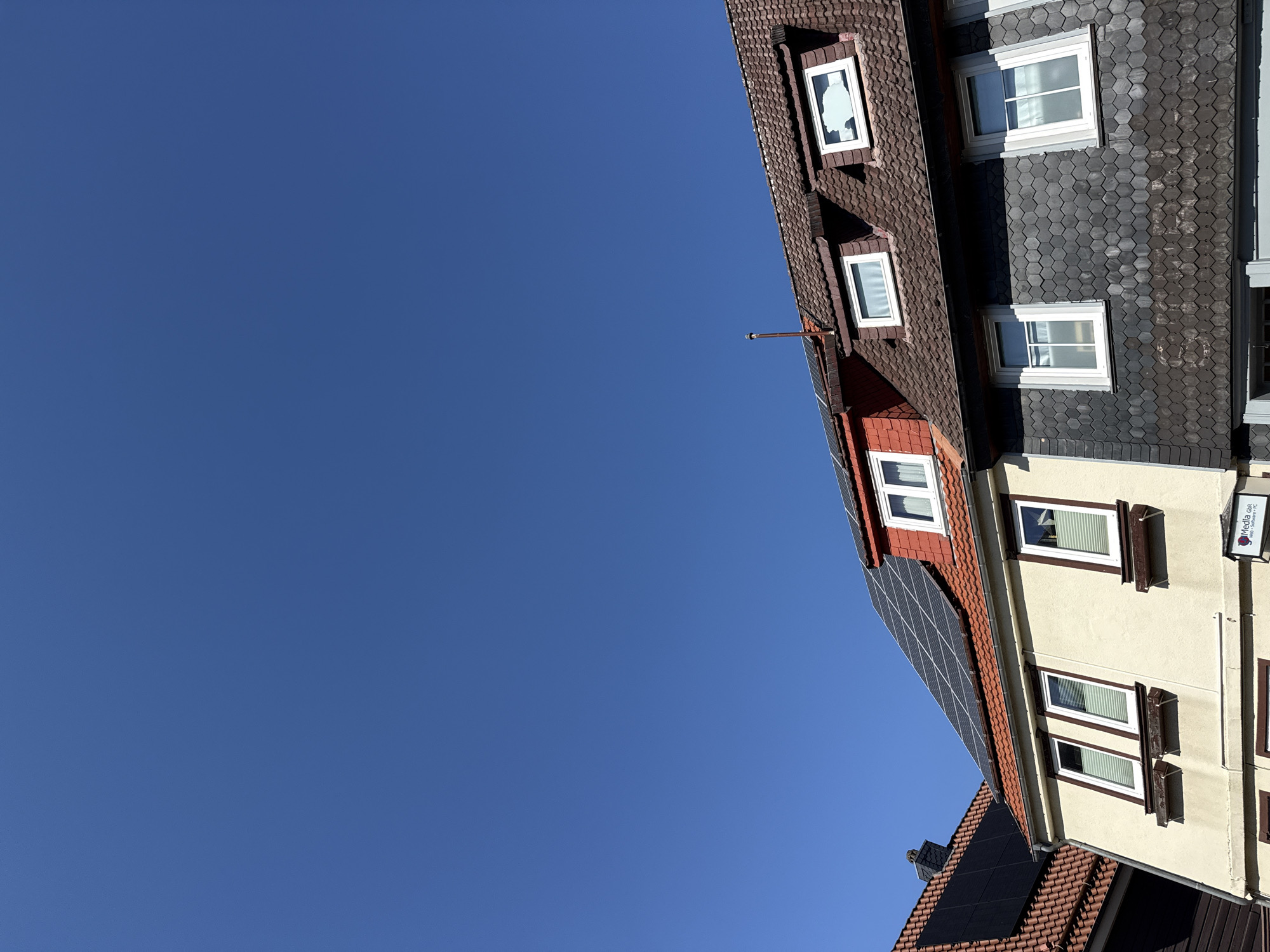 This is a low-angle, eye-level shot of two adjacent buildings under a clear, vibrant blue sky in Herzberg, Germany. The building on the left has a light yellow facade and a terracotta-tiled roof with solar panels. It features three windows on the upper level, each with white frames and blinds. The building on the right has a darker, possibly slate, tiled facade on its lower section and a terracotta-tiled mansard roof with dormer windows. One dormer window on the right has a broken pane. Several dormer windows are visible on the mansard roof, and some also have solar panels on the lower slope. A small chimney protrudes from the right building's roof. A sign is visible on the lower part of the left building, partially obscured, but appears to read "Media GbR..." and includes symbols for "Web + Software + PC." The overall impression is one of urban residential or commercial architecture under bright daylight.