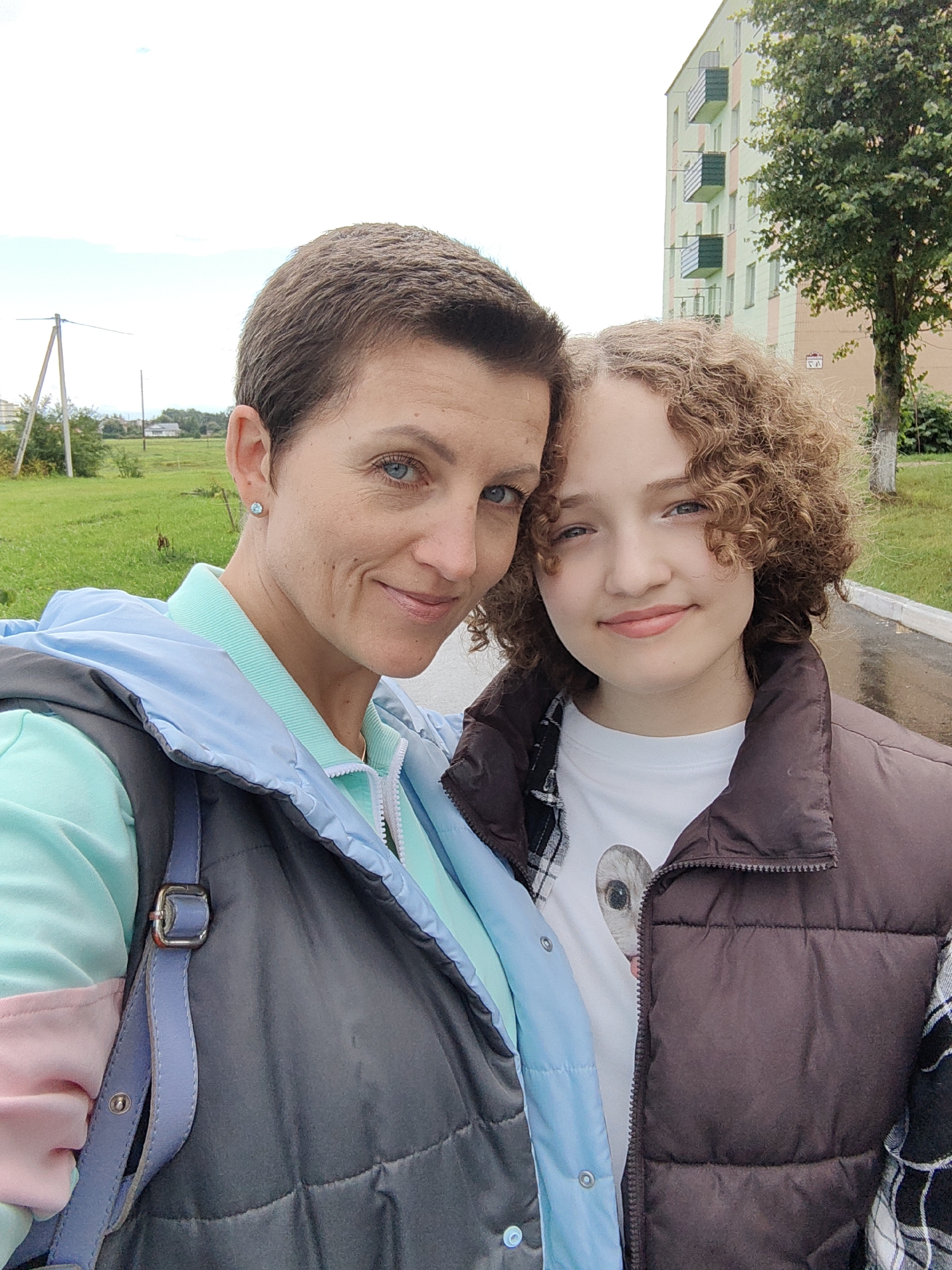 In Myadzyel, Belarus, a woman with short brown hair and blue eyes, wearing a light blue jacket over a mint green shirt and a dark vest, is posing for a selfie with a teenage girl who has curly brown hair and blue eyes. The girl is wearing a white t-shirt with a cat graphic and a brown vest over a plaid shirt. They are standing outdoors with a grassy area, a building, and a tree in the background. The sky is overcast.
