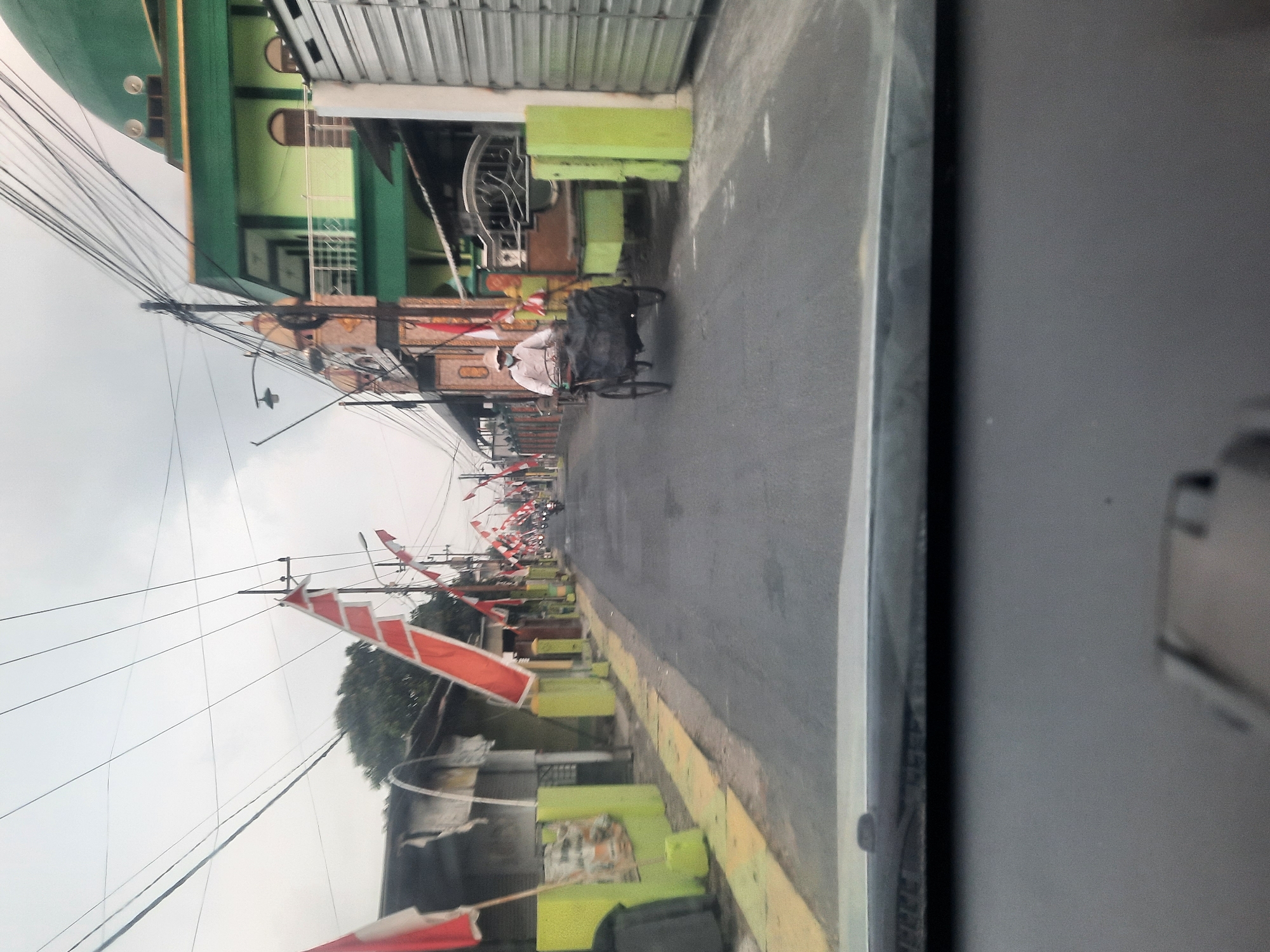The image depicts a street scene in Banjarkemantren, Indonesia, likely during a period of national celebration, as evidenced by numerous red and white flags adorning the street. The perspective is from inside a vehicle, looking forward through the windshield.
In the foreground, the dashboard of a car is visible, with a portion of the windshield wipers and the edge of the windshield frame. The road ahead is paved and asphalted, with a yellow painted curb on the left side.
The street is lined with buildings on both sides. On the left, there are low-rise structures, some with faded paint and what appear to be awnings or open fronts. On the right side of the street, a more prominent building with a green exterior and a domed roof, likely a mosque or religious institution, is visible. This building features balconies with white railings and arched windows. Adjacent to it are more modest buildings and fences.
Several red and white flags are hung along the street, attached to poles and wires, indicating a festive atmosphere. In the middle ground, a person is seen riding a three-wheeled bicycle-rickshaw. The person is wearing a light-colored shirt and a face mask, and the rickshaw is carrying a dark-colored load covered by a tarp. Further down the street, more flags are visible, and there are hints of other vehicles and possibly pedestrians. The sky is overcast, suggesting a cloudy or early morning/late afternoon lighting condition. The overall impression is of a typical urban or semi-urban street in Indonesia decorated for a special occasion.