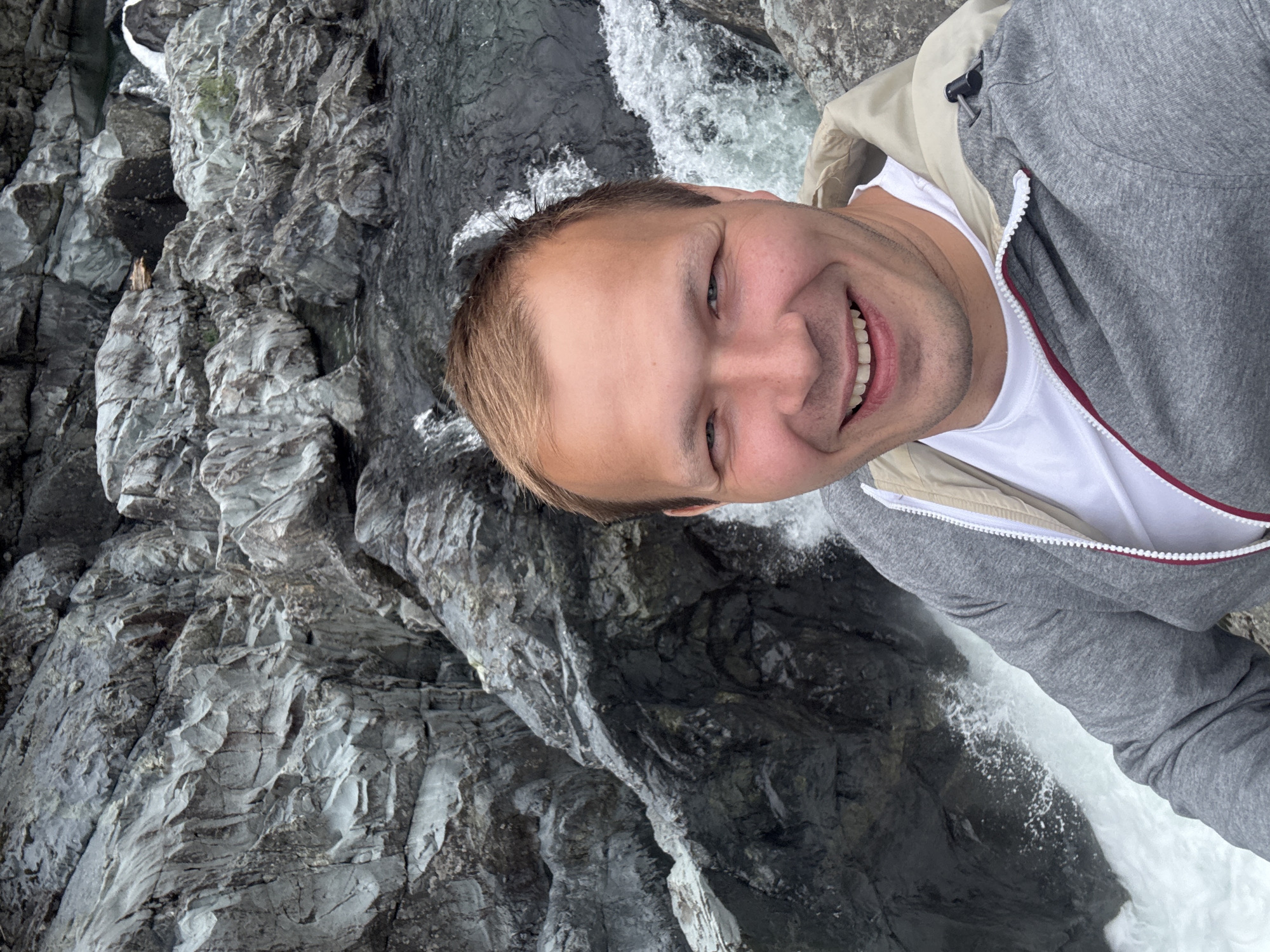 A fair-skinned man with short, light brown hair smiles at the camera, positioned at an angle. He is wearing a white t-shirt and a gray hooded sweatshirt with a beige lining. The background consists of a gray rock face and a rushing waterfall. The location is Area C in Long Beach, Canada.