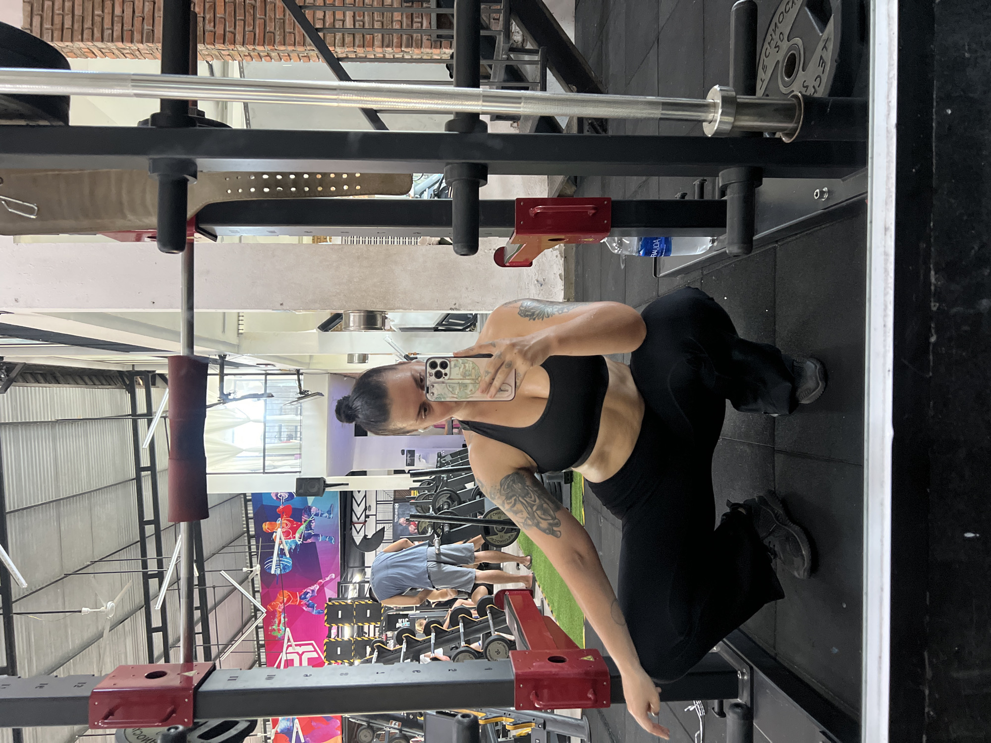 The image shows a woman taking a mirror selfie in a gym, located in Hội An Ward, Vietnam. She is wearing a black sports bra and black leggings, with her hair pulled up in a bun. She is in a squatting position in front of a squat rack. In the background, there are other gym equipment, including weight plates and machines, and other people working out. The gym appears to be spacious with a high ceiling and industrial-style lighting. The overall environment suggests a functional fitness facility.