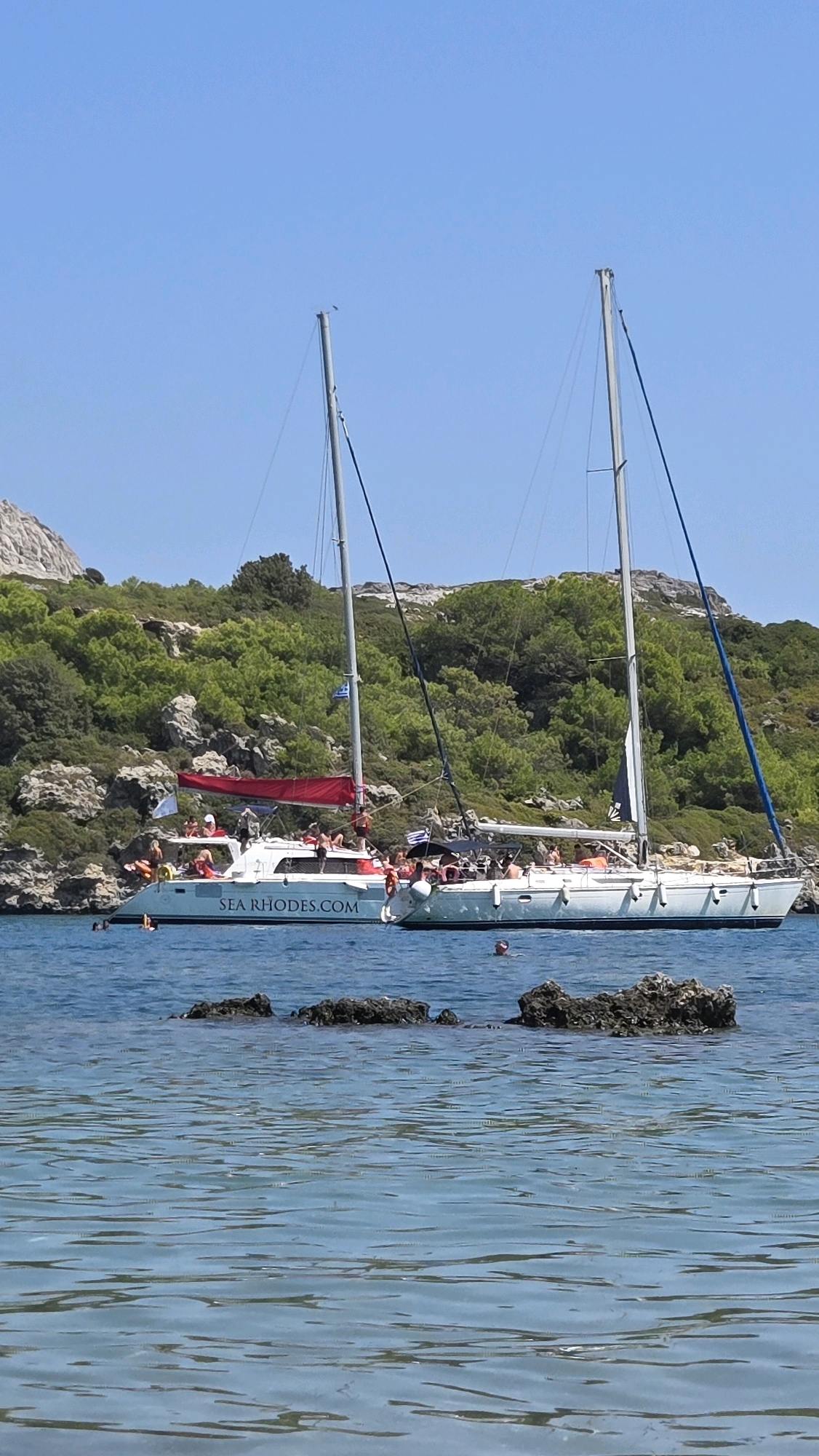 The image depicts a bright, sunny day at sea with a rocky, tree-covered coastline in the background. Two sailboats, a catamaran and a monohull, are anchored in the clear blue water. The catamaran, with the text "SEA RHODES.COM" on its side, is white with a red sail. It is carrying numerous passengers enjoying the scenery and water. The monohull sailboat is to the right of the catamaran, also with passengers on board. Several individuals are swimming and wading in the shallow water around submerged rocks in the foreground. The overall scene suggests a recreational boat trip or excursion in the waters near Rhodes, Greece.