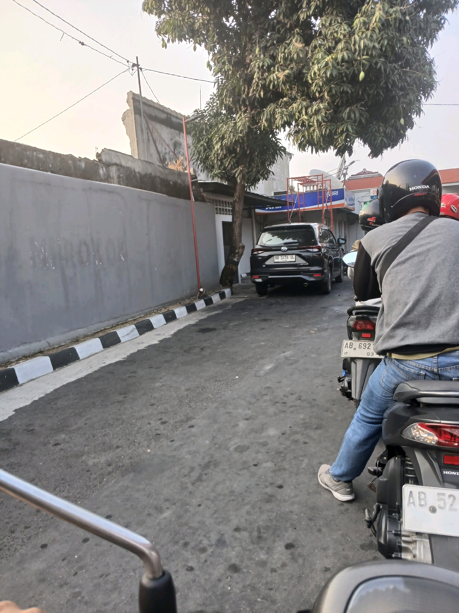 A street scene in Depok, Indonesia, captured from a motorcycle's perspective. The road is paved and appears to be a public street with a grey wall on the left side, featuring a black and white striped curb. A black SUV is parked on the right side of the road, with its license plate reading "AB 1476 UL". Further ahead, there are two individuals on motorcycles. The person in the foreground, whose back is to the camera, is wearing a grey shirt and blue jeans, and is seated on a motorcycle with the license plate "AB 692". Another motorcycle is visible in front of them, with a rider wearing a red helmet. A large tree with mangoes is prominently featured on the right side of the image. In the background, there are buildings and utility wires. The sky is overcast.