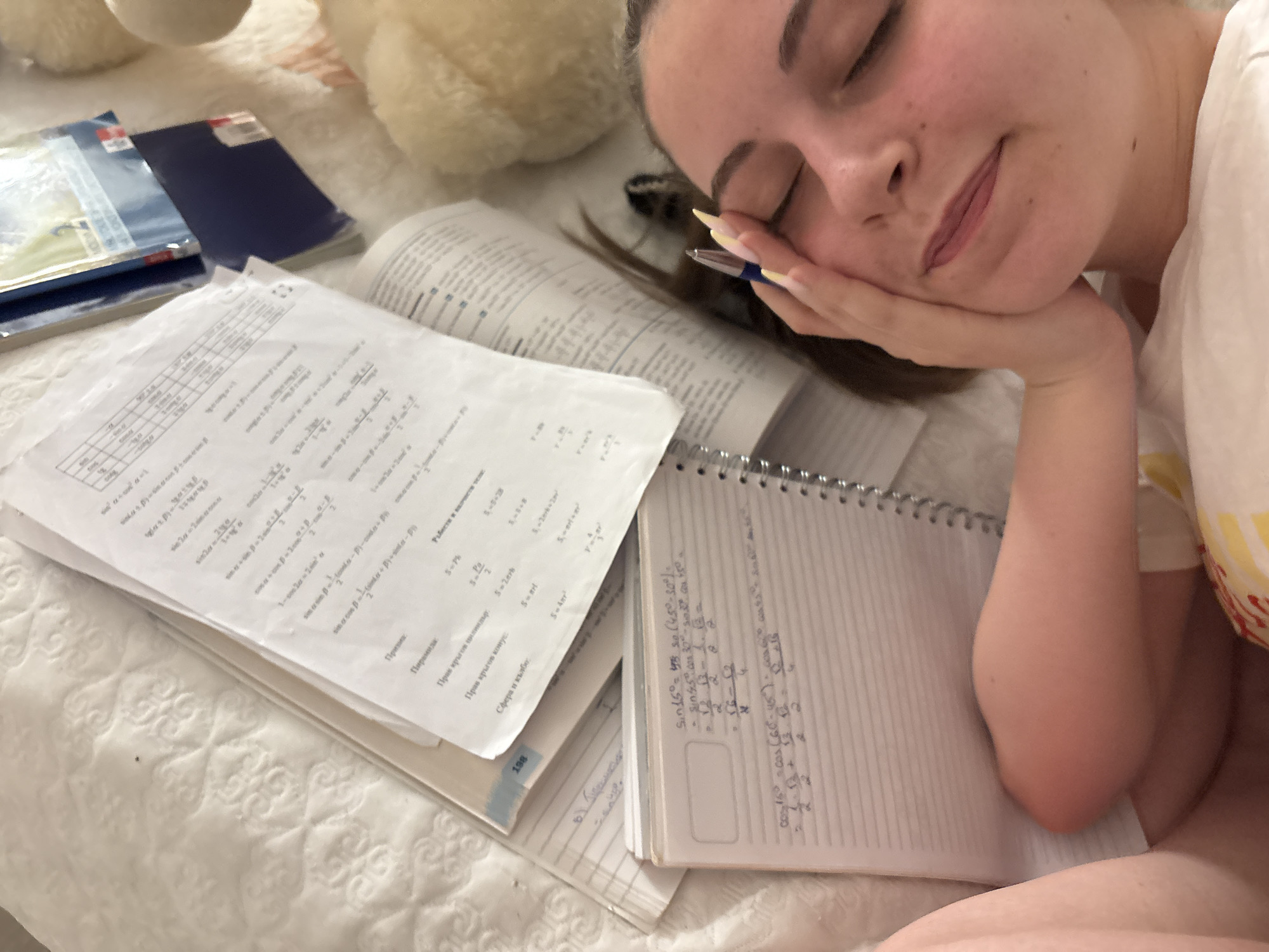 A young woman lies with her eyes closed on a white patterned surface, possibly a bed, surrounded by study materials. A notebook filled with mathematical calculations lies open beside her. A textbook and loose-leaf papers covered with formulas and written notes are scattered nearby. The location is not Bulgarovo, Bulgaria.