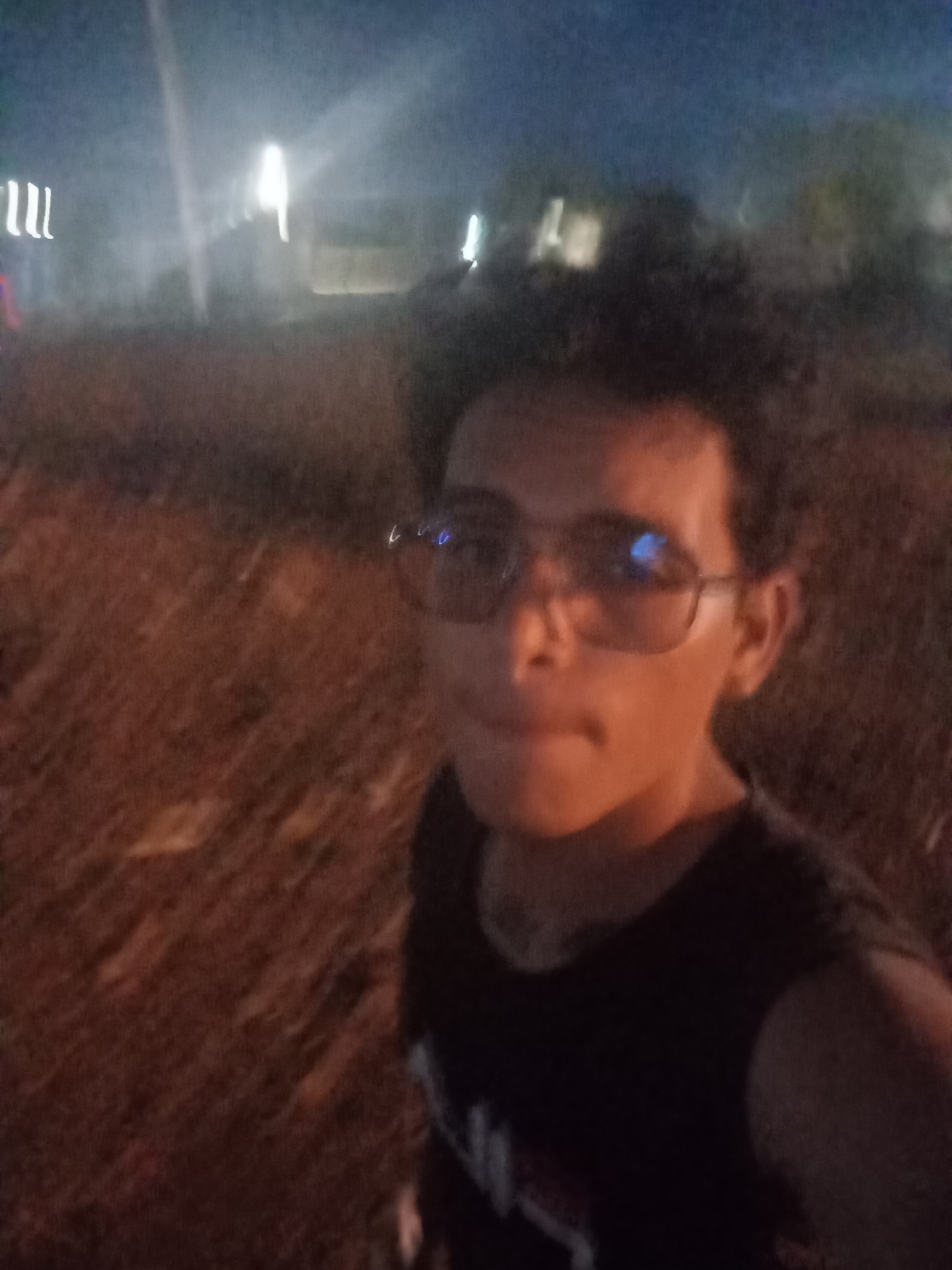 The image is a blurry, low-light, close-up selfie of a young person wearing glasses and a dark t-shirt. The background suggests an outdoor setting at night, with indistinct lights and foliage. The person's face is illuminated by warm artificial light, casting shadows and highlighting their features. The glasses reflect blue light, possibly from a screen or another light source. The overall scene is somewhat chaotic due to the low resolution and motion blur. The image does not contain enough discernible information to confirm a specific location within Altagracia, Venezuela.