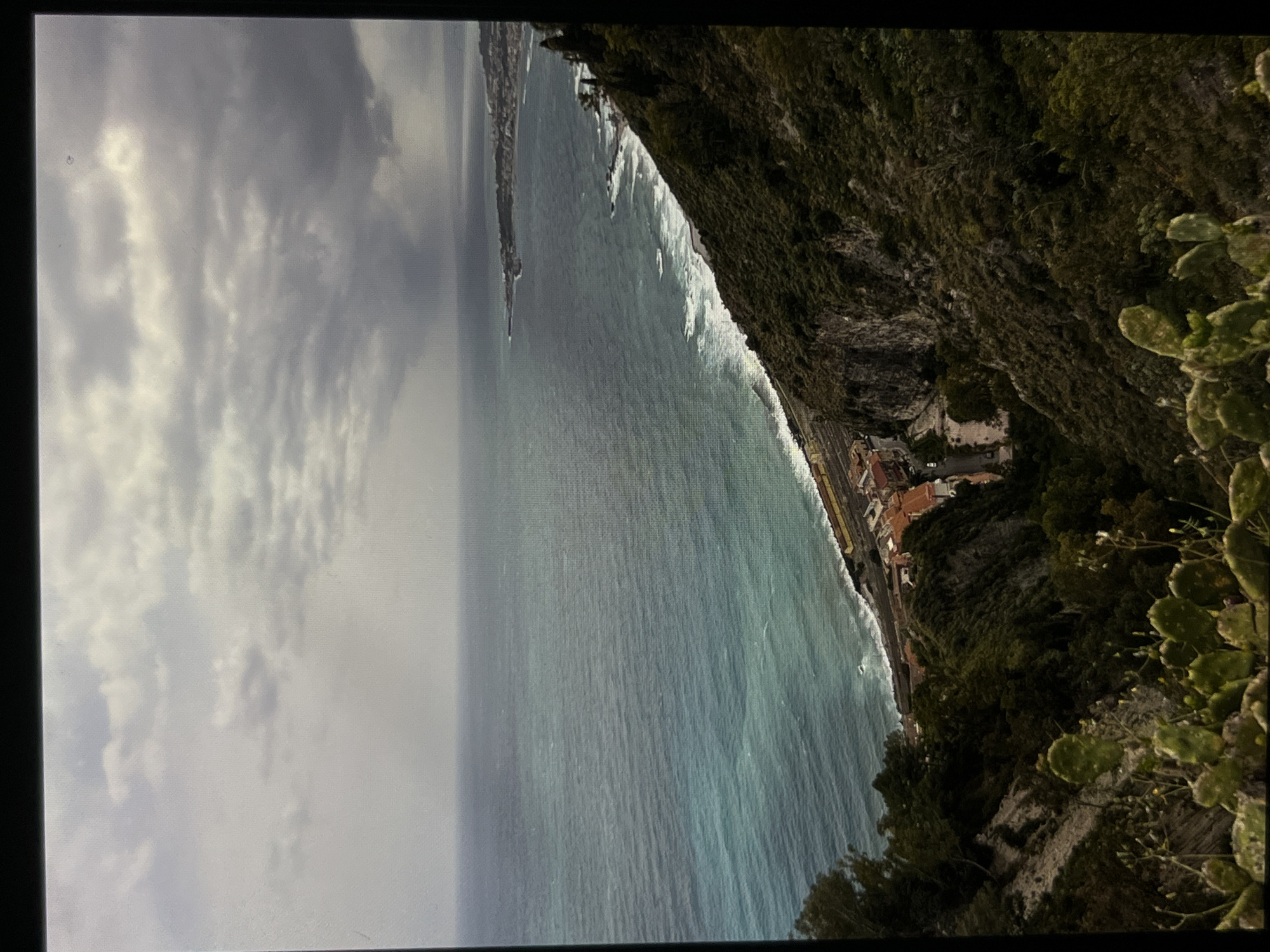 The image is an aerial view of a coastal landscape with a city nestled along the shore. The sea is a vibrant turquoise, with white waves crashing onto the rocky coastline. A road and railway line run parallel to the sea, with a cluster of red-roofed buildings adjacent to them. The land slopes upwards from the coast, covered in dense green vegetation and rocky outcrops. In the foreground, prickly pear cacti are visible, adding to the natural scenery. The sky is overcast with heavy grey clouds, suggesting a moody atmosphere. The overall scene depicts a rugged and scenic coastal area, likely in a Mediterranean region, with human settlements integrated into the natural environment.