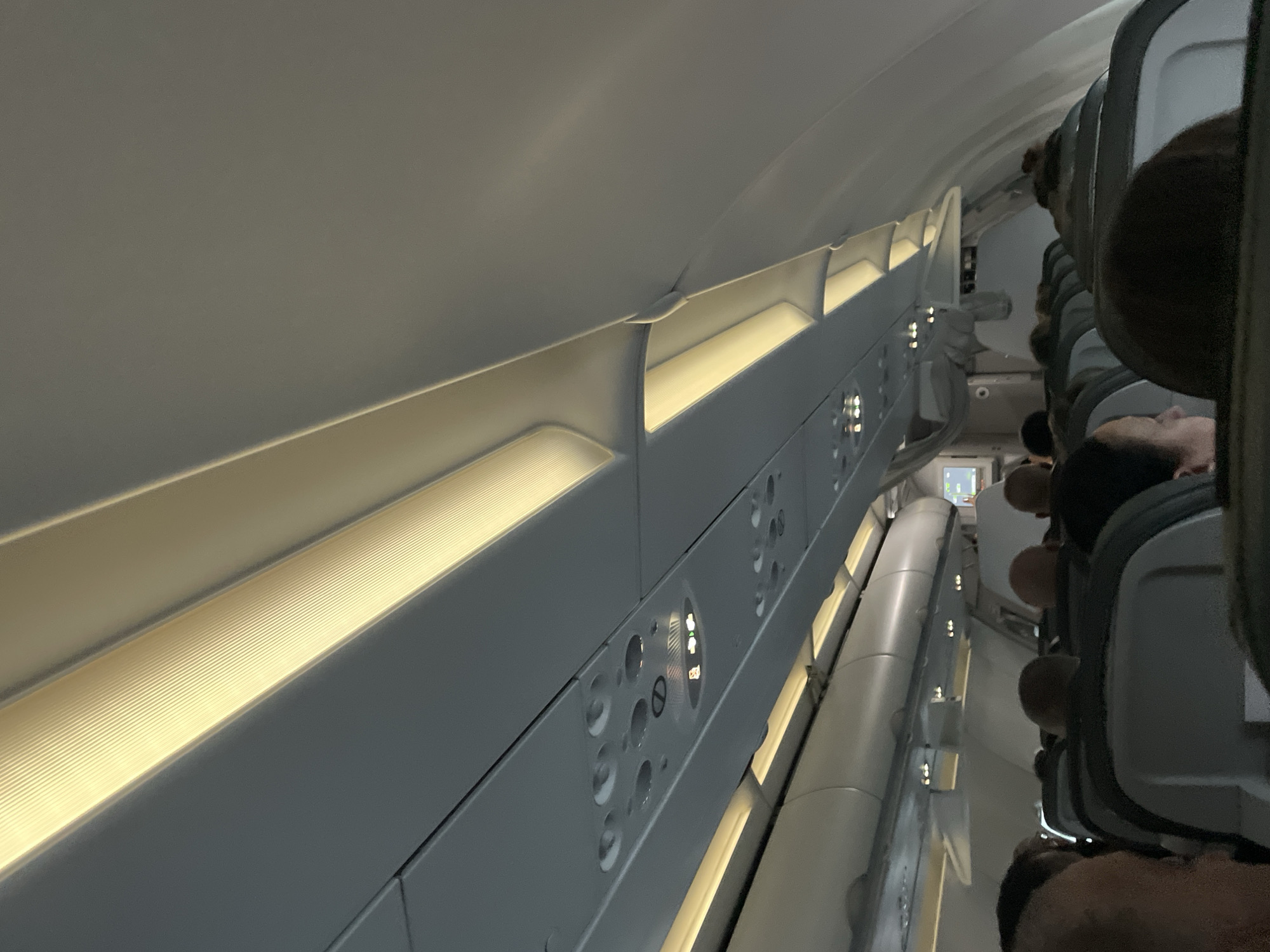 The image depicts the interior of a commercial airplane cabin. Passengers are seated in rows of seats. Overhead compartments are visible along the upper part of the cabin interior. The lighting inside the aircraft is bright. The location is not discernible from the visual information provided.