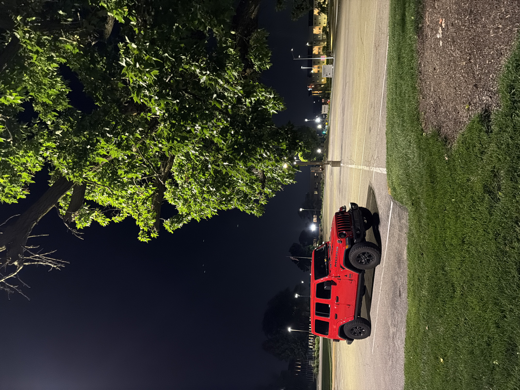 In an outdoor parking lot at night in Maryland Heights, Missouri, a red four-door Jeep Wrangler is parked. The vehicle is positioned in the foreground, angled slightly towards the viewer, with its front end visible. The Jeep has black tires and a black grille. To the right of the Jeep, a large tree with green leaves dominates the frame, its branches extending overhead and partially obscuring the dark night sky. The parking lot is paved and marked with white lines. In the background, streetlights illuminate the lot, and several buildings with lighted windows are visible. A few faint white specks, possibly birds or insects, are scattered in the sky. The ground in the lower portion of the image is covered with green grass.