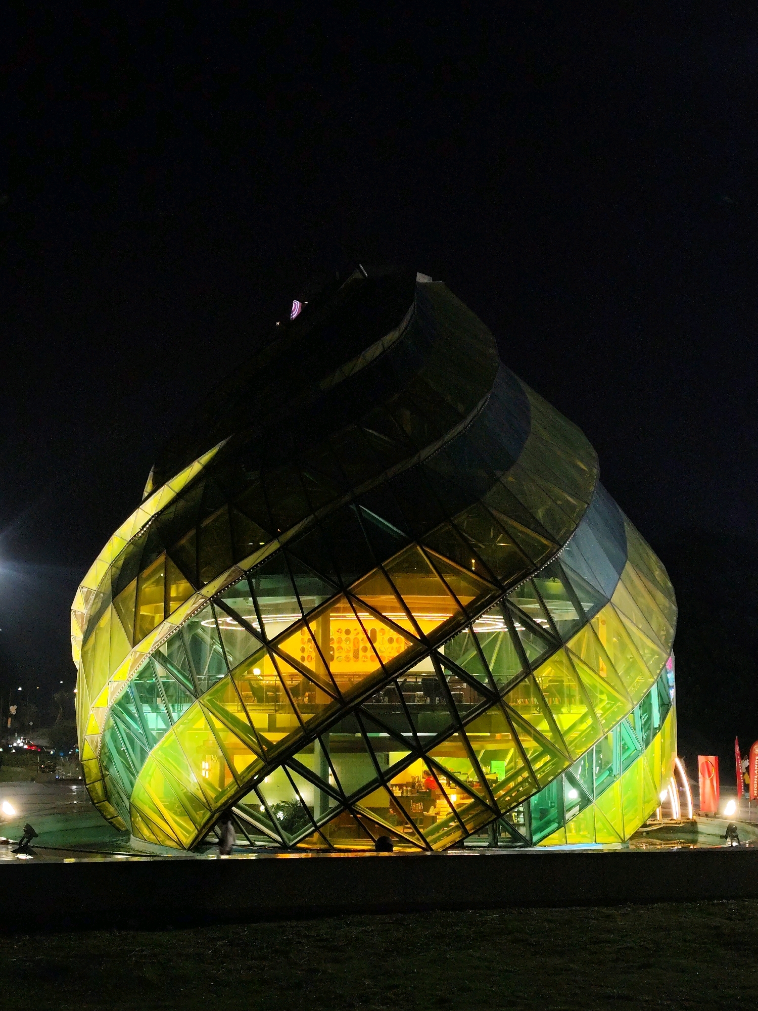 The image shows a modern, geodesic-domed building at night in Da Lat, Vietnam. The building's exterior is covered in diamond-shaped glass panels that are illuminated with a gradient of yellow and green lights, giving it a striking, almost iridescent appearance. The structure appears to be multi-story, with visible interior lighting that highlights the internal architecture and hints at activity within. In the foreground, there is a dark, grassy area, and in the background, the dark night sky is visible with distant city lights. There are a few figures of people present, appearing as silhouettes against the illuminated building, suggesting the building is a public space or a commercial establishment that is open at night. The overall impression is one of striking contemporary architecture set against the backdrop of a darkened urban environment.