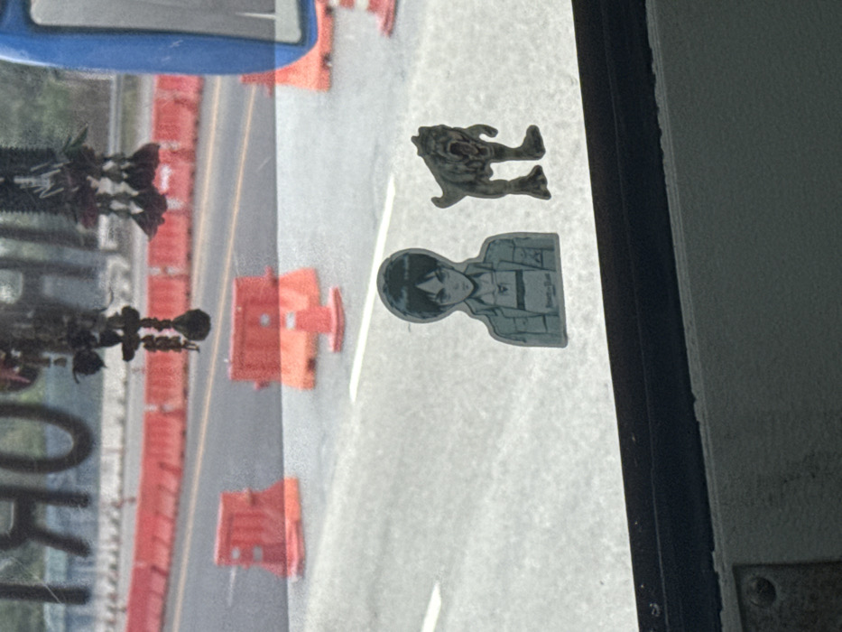 The image presents a view from inside a vehicle, looking outward through a window onto a road in Bang Sao Thong Subdistrict, Thailand. Two stickers are adhered to the lower section of the window: one is a light-toned depiction of an anime character, identified as Eren Yeager, facing forward with a neutral expression; the other, positioned to its right, is a three-dimensional figure resembling a roaring bipedal dinosaur or monster. Reflections are visible in the upper part of the window, including mirrored text partially legible as "IMPORT" and dark, ornate hanging items, possibly vehicle decorations. A portion of a blue vehicle component, likely a side mirror housing, is visible on the extreme right. The exterior environment consists of an asphalt road with visible white lane markings. Numerous orange plastic road barriers and red traffic cones are arranged along the road, indicating a controlled or work zone.