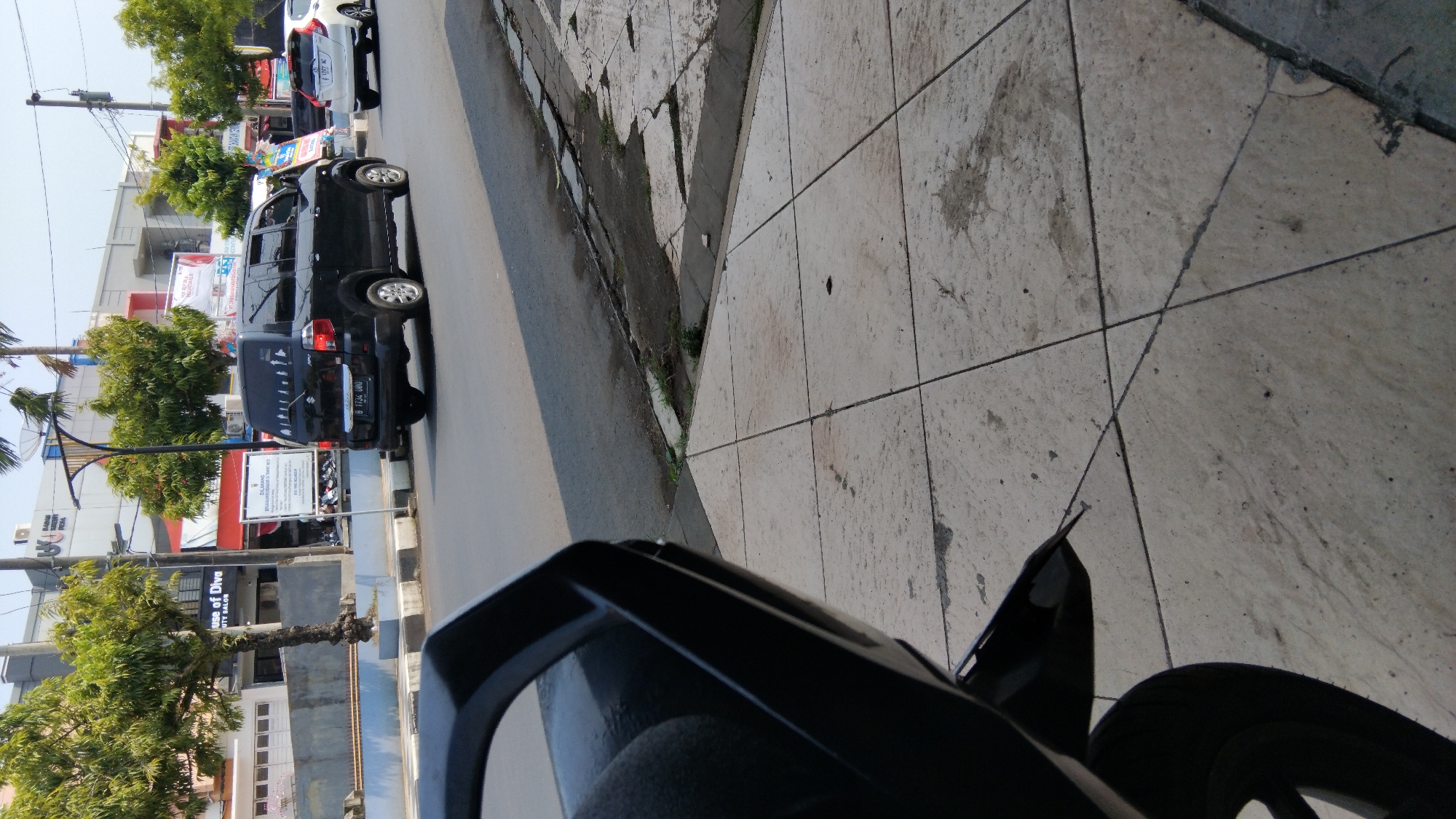This image captures a street scene in Batang, Indonesia. A black minivan is traveling on the road, with its license plate visible as "B 1734 UUD". To the right of the minivan, a white car is also on the road. The foreground of the image shows a tiled sidewalk and a portion of a black motorcycle. In the background, there are several trees and buildings. One building has signage indicating "House of Diva Beauty Salon". There are also street signs and utility poles visible. The sky is clear and bright, suggesting it is daytime.