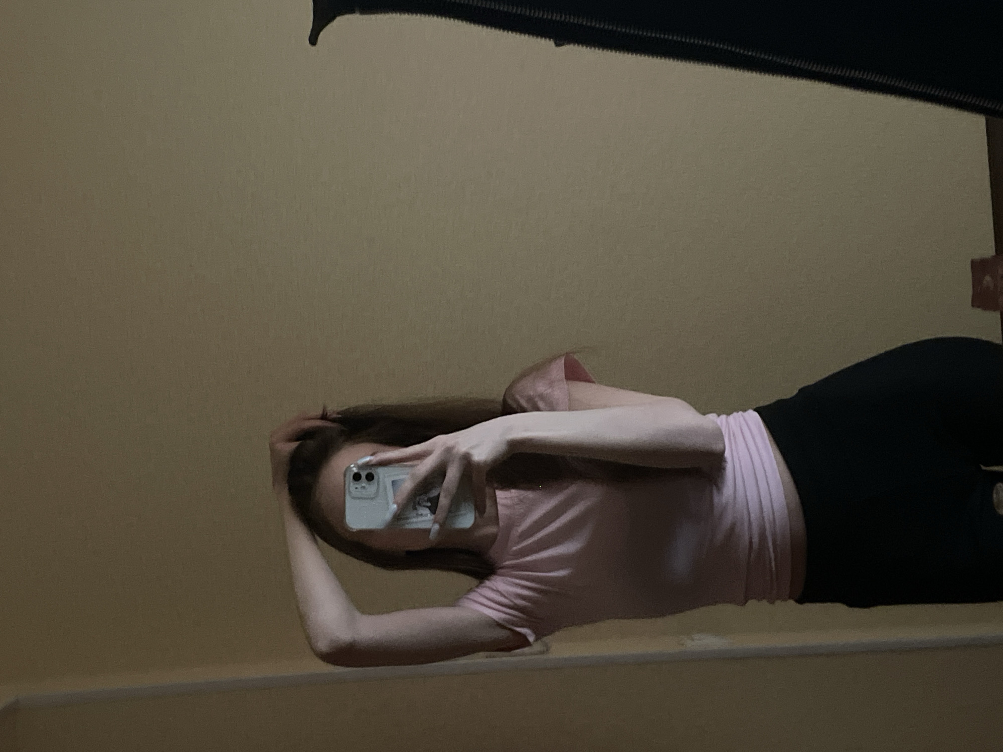A person is taking a mirror selfie in what appears to be an indoor environment with yellowish walls. The person is wearing a light pink t-shirt and black leggings. The phone partially obscures their face. The image contains an edge of a mirror with a black frame on top and a white edge on the bottom, presumably from a countertop or shelf. The location in the city of Ufa, Russia, cannot be determined from the visual data.