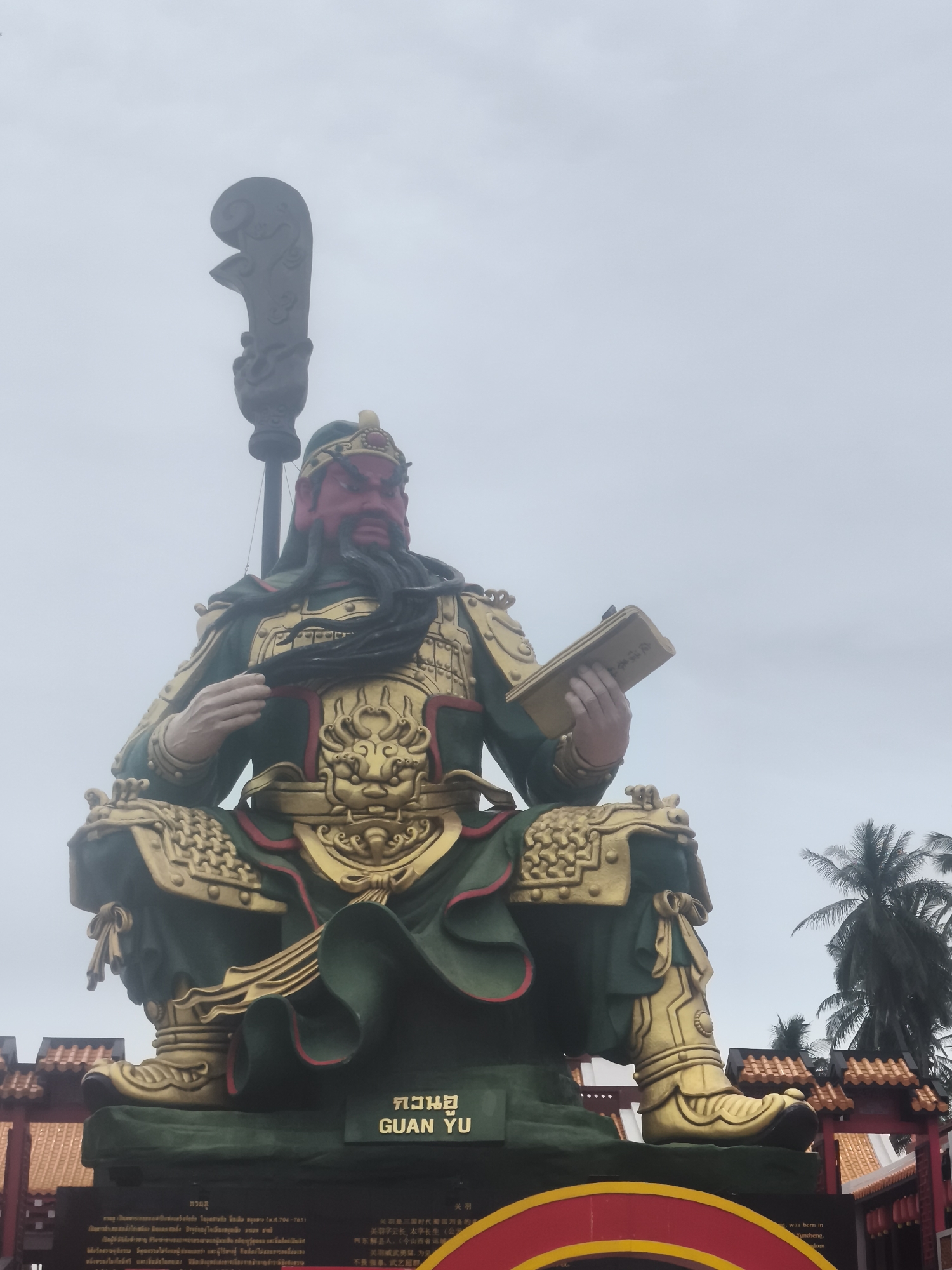 A large, imposing statue of Guan Yu, a Chinese general, is depicted sitting cross-legged against a pale sky in Baan Hua Thanon, Thailand. The statue is predominantly green with ornate gold accents on its armor, helmet, boots, and throne. Guan Yu's face is red, with a long black beard and intense eyes. He is holding a golden book in his right hand, and a large, decorative weapon is positioned vertically behind his left shoulder. The base of the statue has Thai and Chinese inscriptions. In the background, palm trees are visible, and parts of a temple structure with orange tiled roofs can be seen to the left and right of the statue. The overall atmosphere is solemn and grand.