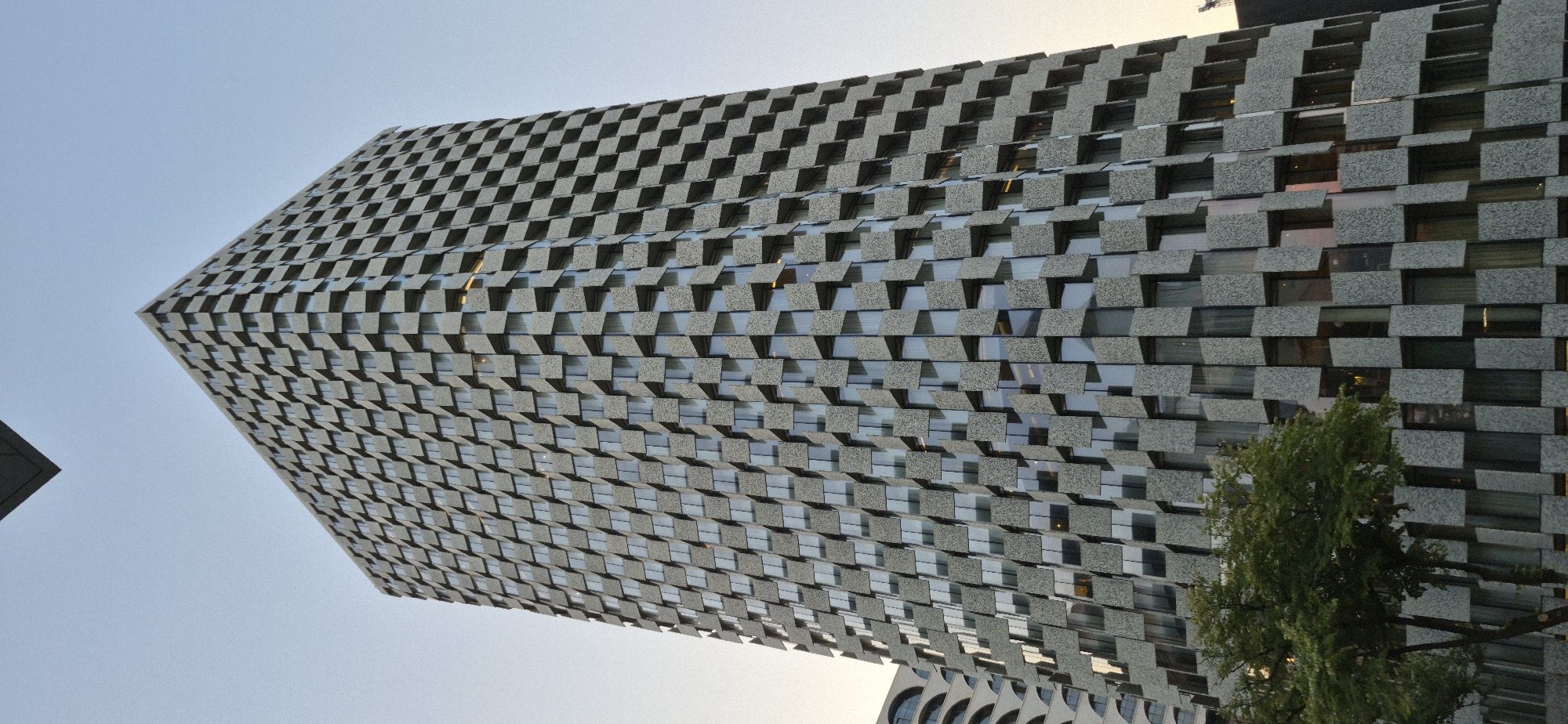 The image shows the "Tirana International Hotel and Business Center," a prominent skyscraper located in Tirana, Albania. The building features a distinctive facade composed of alternating glass panels and concrete blocks, creating a dynamic, tessellated pattern. The concrete blocks are arranged in a staggered, almost three-dimensional manner, giving the impression of protruding squares. The building's exterior is a neutral gray, with the glass windows reflecting the sky and surrounding environment.
A mature tree with lush green foliage stands in the foreground, partially obscuring the lower floors of the building. The tree's branches reach upwards, complementing the vertical lines of the skyscraper. In the background, another building with arched windows can be seen on the left, suggesting an urban setting with varied architectural styles. The sky above is a pale, muted blue, indicative of either early morning or late evening. There are no people visible in the image. The overall scene is static, focusing on the architectural grandeur of the hotel and its surrounding environment.