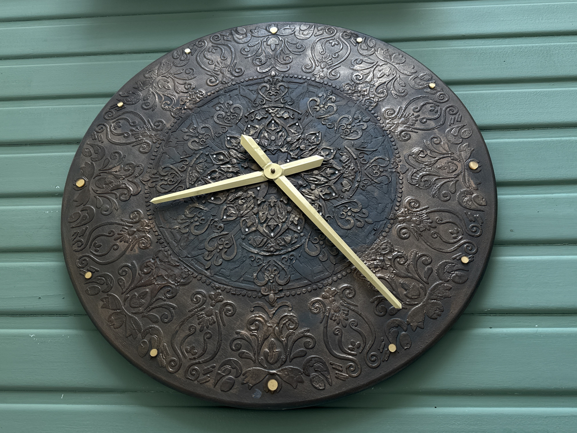 A round, ornate clock with golden hands hangs on a teal wooden paneled wall. The clock face is dark with raised, intricate patterns in gold and bronze. There are small golden dots around the perimeter of the clock face, serving as markers for hours. The clock hands are pointing to approximately 11:20. The wall is painted a deep teal color and has a vertical, paneled design. The lighting is soft, casting gentle shadows. The image does not contain any people or specific identifiable locations within the city of Kolomna, Russia.