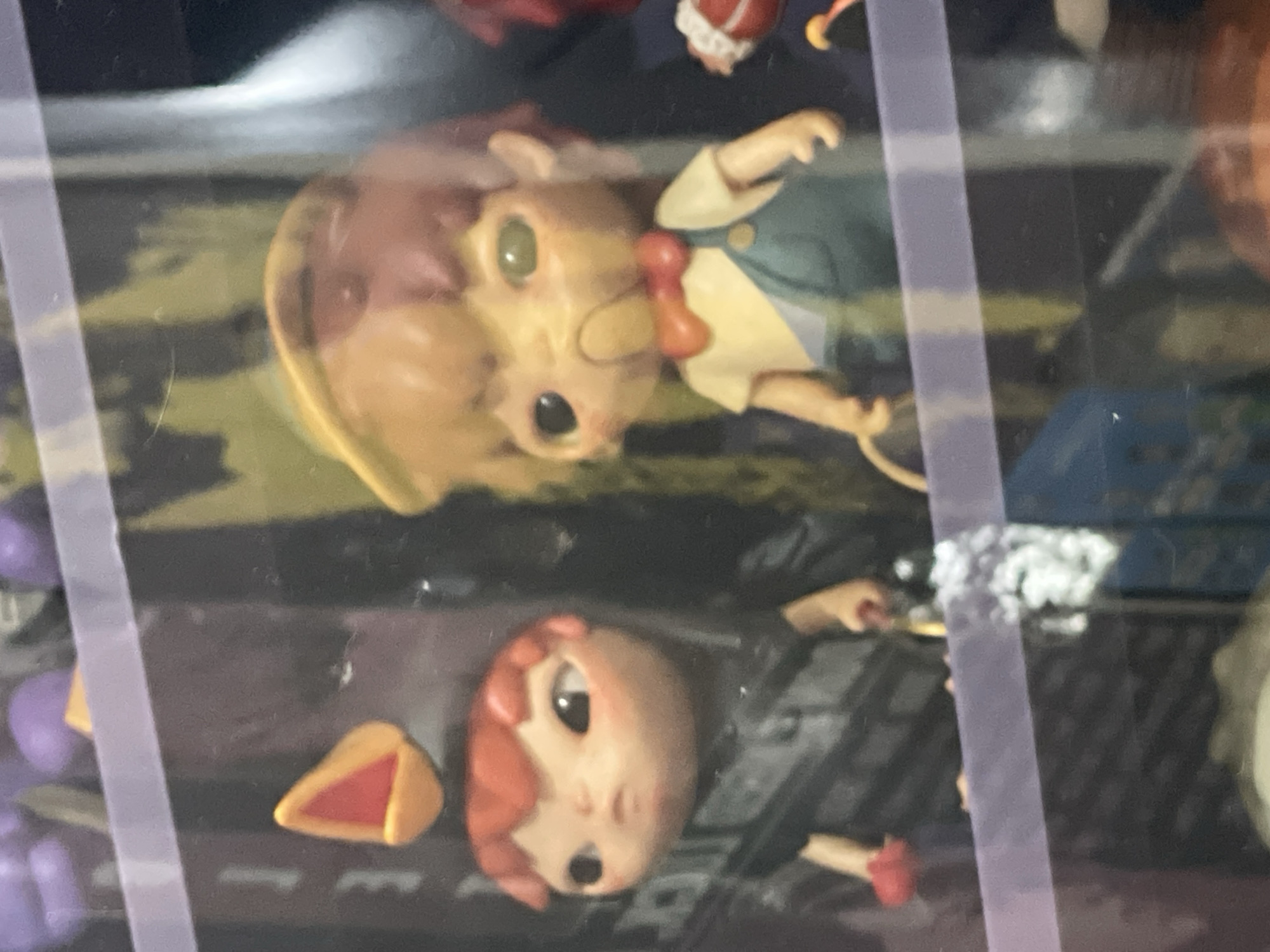 The image displays a close-up view of several collectible figurines arranged in what appears to be a display case. In the foreground, on the left, is a figurine with red hair and a dark outfit, holding a red object in its left hand. To its right, slightly further back, is another figurine wearing a yellow shirt and blue overalls, with a red bow tie. This figurine has a distinct yellow mask-like feature over its mouth and nose and a yellow hat. The background shows blurry purple and green shapes, possibly other figurines or decorative elements. A translucent purple horizontal band runs across the middle of the image. The lighting suggests an indoor setting. There is no information in the image that indicates the location is Denpasar, Indonesia.