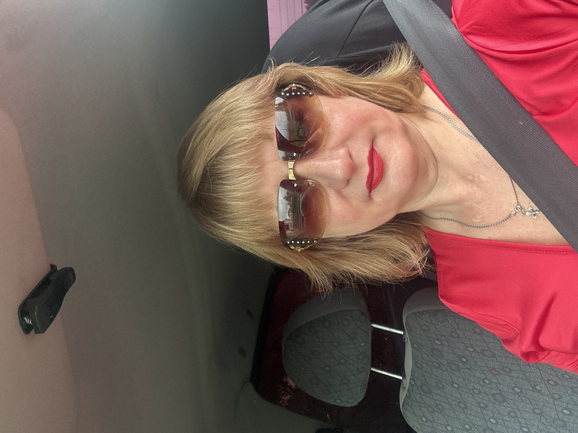 A selfie of a middle-aged Caucasian woman with blonde hair and sunglasses is taken inside a vehicle. She is wearing a red shirt and a silver necklace with a cross pendant. The woman is looking directly at the camera. The interior of the car is visible, including a gray patterned seat and a seatbelt. A black object is clipped to the roof of the car. The background is blurred, but appears to be outdoors, possibly in a rural area. The image is undated and unsourced. The location of Tugulym, Russia, is not discernible from the image itself.