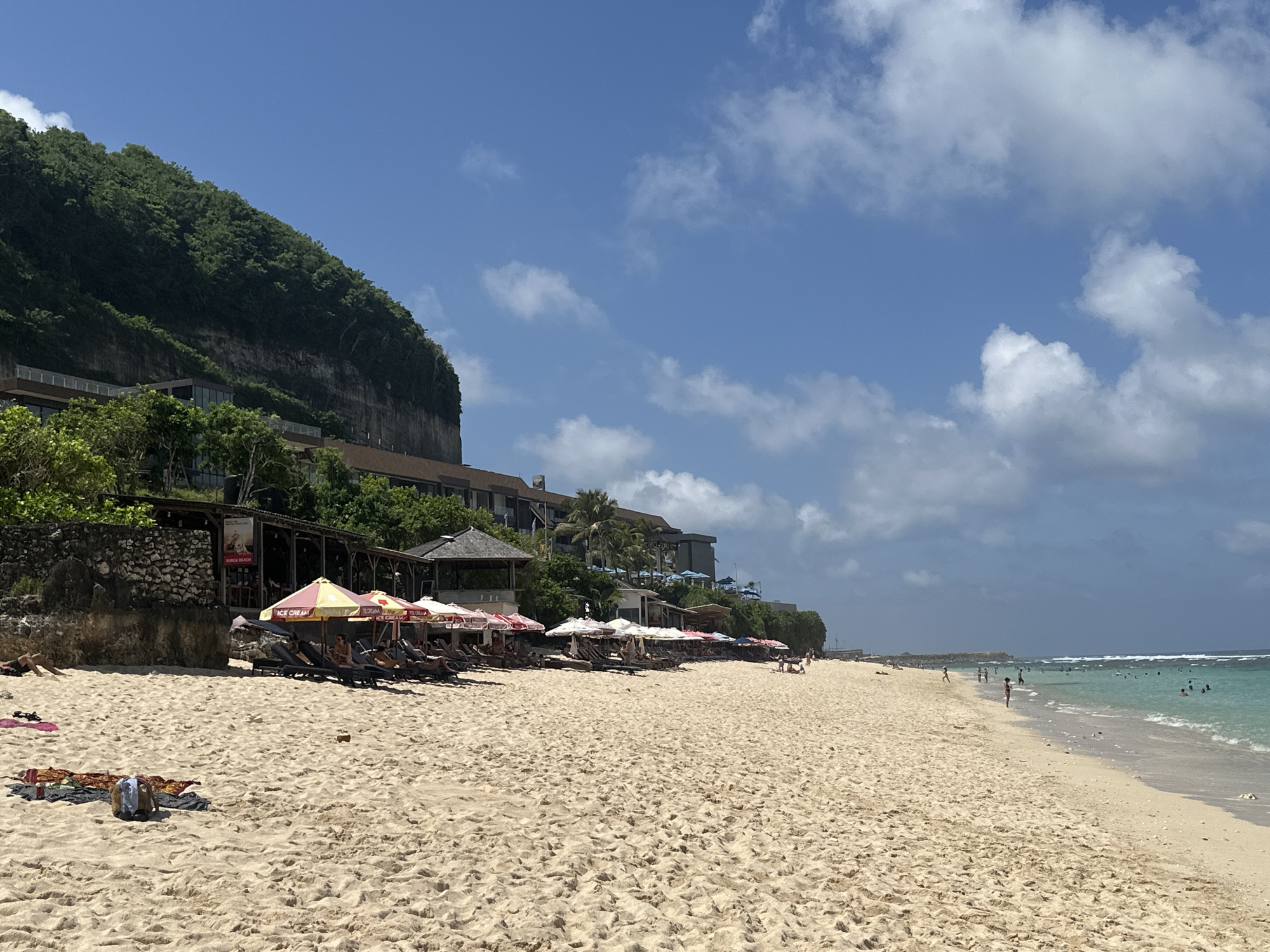 The image depicts a beach scene in Kuta Selatan, Indonesia. In the foreground is a sandy beach populated with people, some lying on towels or sunbathing, and others swimming in the ocean. A row of beach umbrellas and lounge chairs lines the edge of the beach near a series of buildings and structures. Above the buildings is a steep, lush cliff covered in green vegetation. The sky is blue with scattered white clouds. The ocean is visible in the background, with several individuals swimming or standing in the water. The overall scene suggests a sunny day at a beach resort.