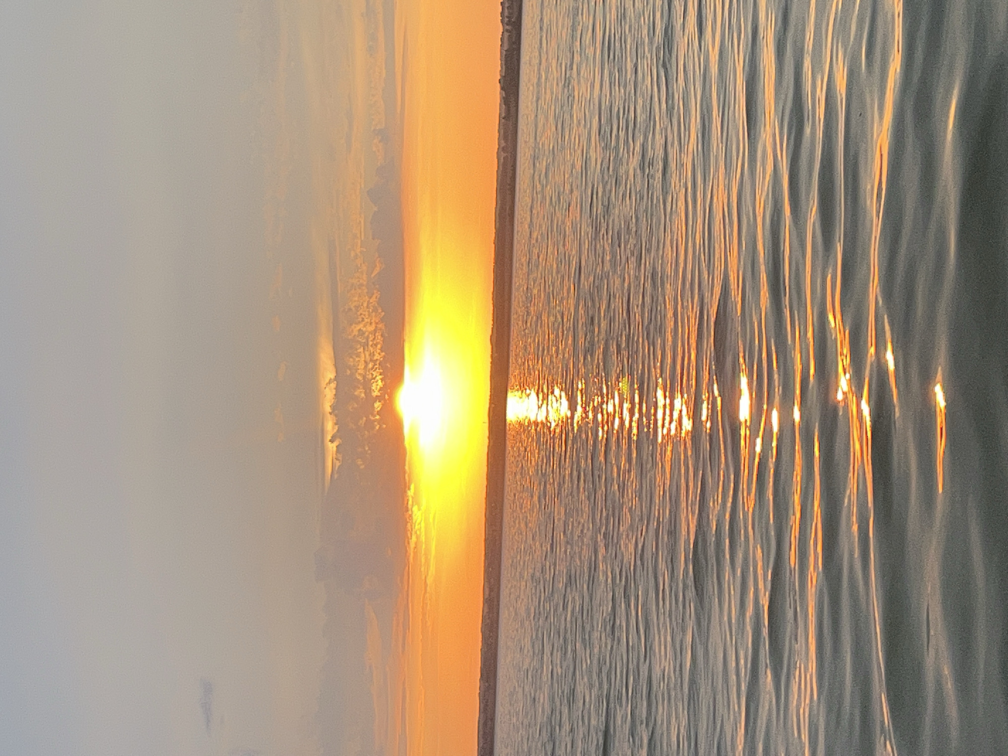 The image depicts a serene sunset over the water. The sun, a bright, intense yellow-orange orb, is positioned low on the horizon, partially obscured by wispy clouds. Its reflection creates a shimmering path of golden light across the rippling surface of the water. The sky transitions from a pale blue at the top to vibrant shades of orange and yellow near the horizon, with scattered clouds catching the warm hues of the setting sun. The water itself appears calm, with gentle waves creating subtle textures and reflecting the colors of the sky. The distant shoreline is visible as a dark, blurry line, suggesting a landmass beyond the water. The scene conveys a sense of tranquility and natural beauty. The location is identified as Beregovoe, Russia.