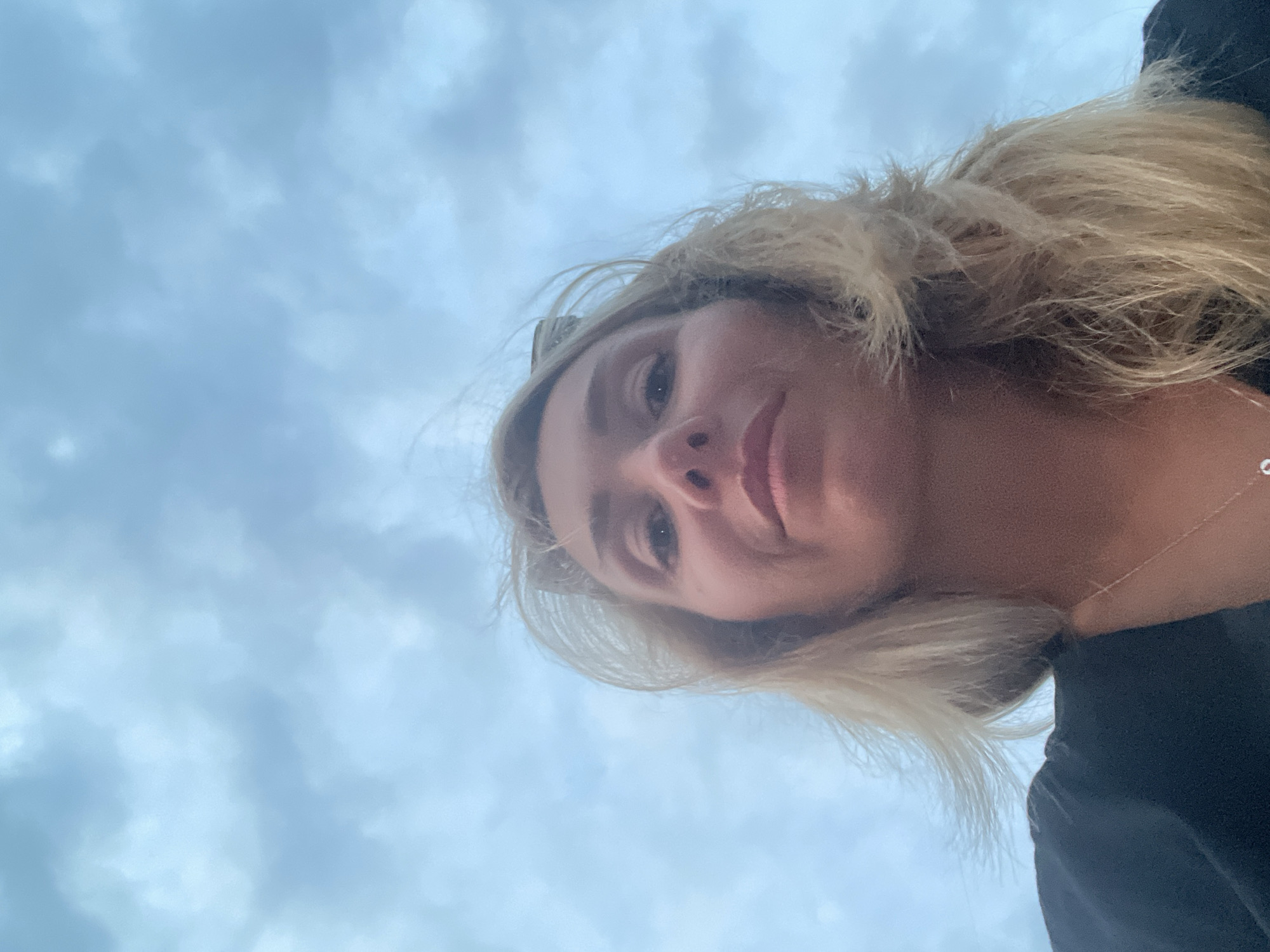 A low-angle, medium shot shows a woman with blonde hair from the chest up, looking at the camera. The woman is wearing a black top and a silver necklace. The background is a cloudy sky with a pale blue hue. The shot appears to be a selfie taken outdoors, possibly in a city given the context of the prompt.