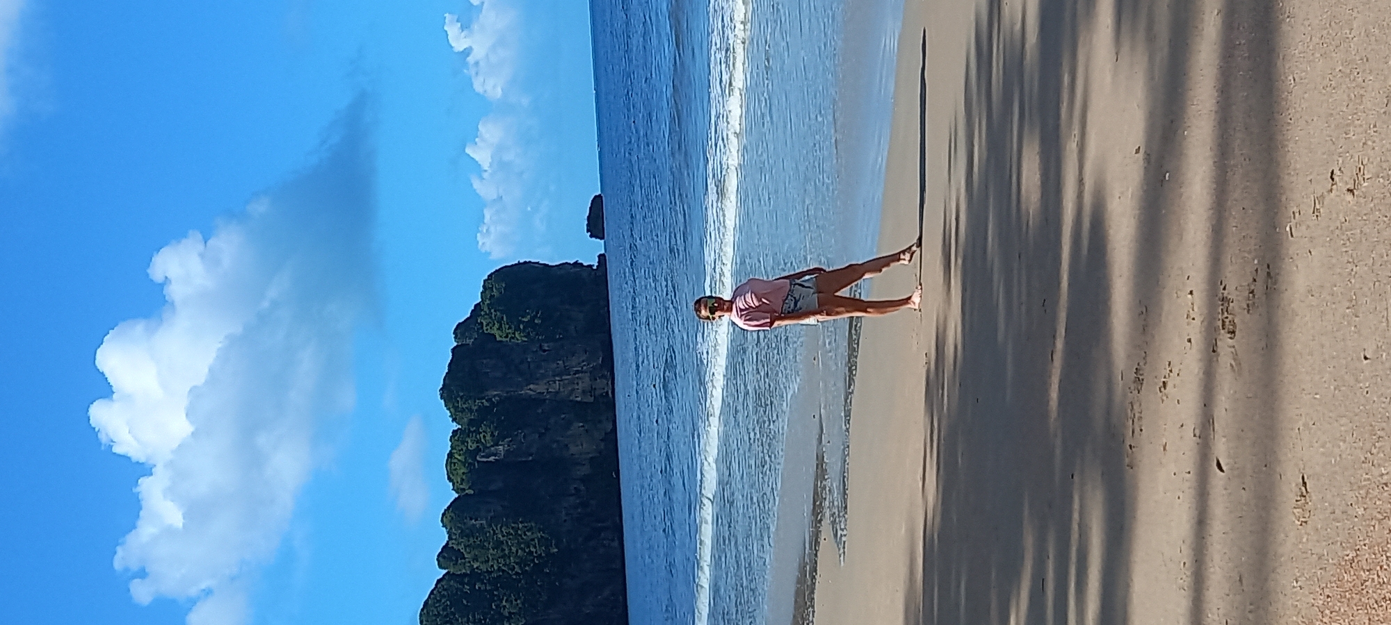 The image depicts a beach in Ban Ao Nang, Thailand. The scene shows a woman in a pink shirt and denim shorts walking along the shoreline. The sea laps at the shore nearby, creating a line of foam. Lush green islands stand prominently in the background against a blue sky with scattered white clouds. Shadows from foliage fall on the sand.