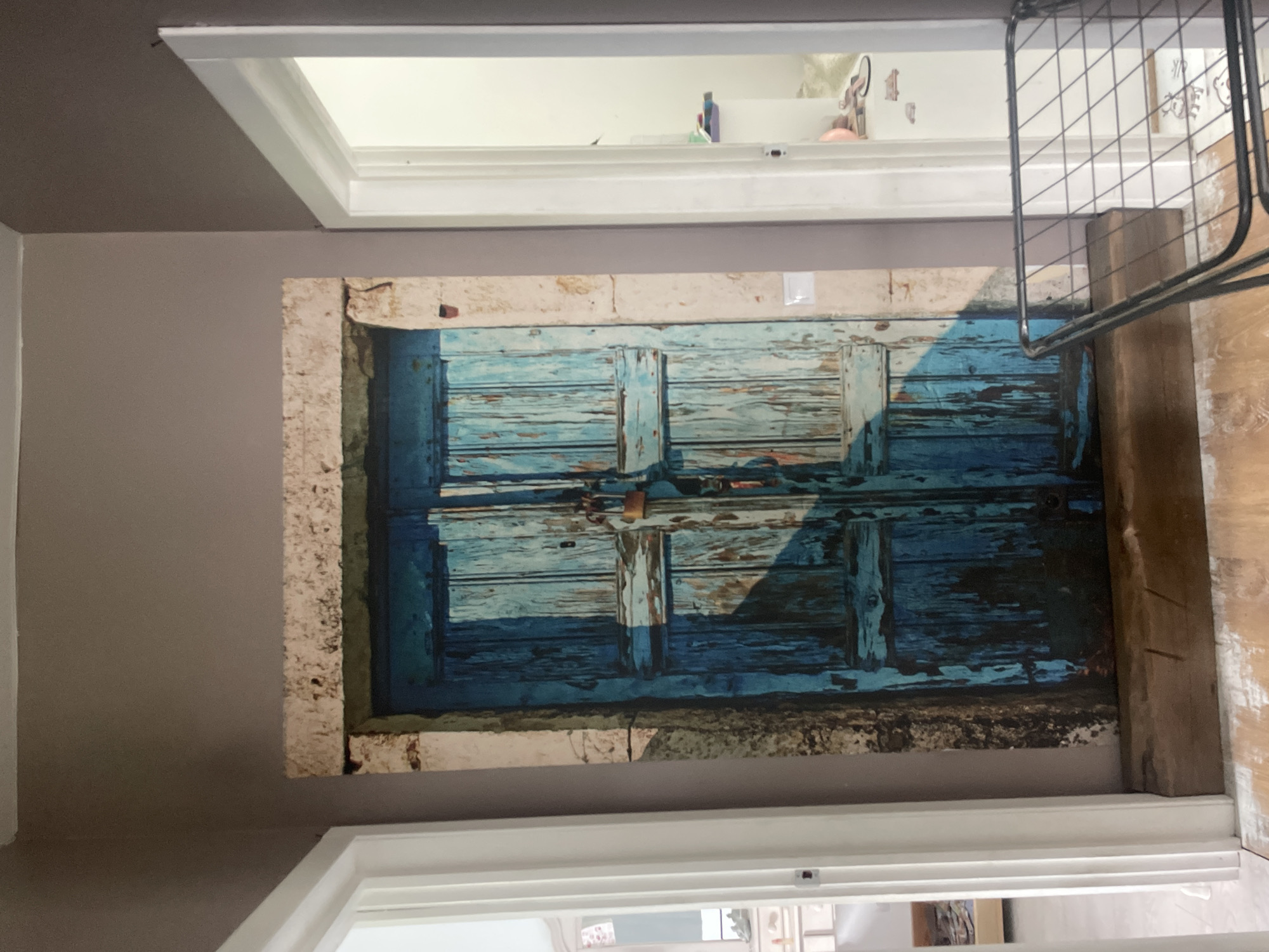The image shows a hallway with a large mural of a weathered blue wooden door. The door appears to be set into an old stone frame. To the left of the mural is a white doorway leading to another room, partially visible. To the right of the mural is another white doorway, also partially visible, showing a brightly lit room with shelves and some objects on them, including what appear to be shoes and a pink object. In the foreground, a black wire rack is placed on the wooden floor, partially obscuring the bottom of the mural and the floor area. The walls of the hallway are painted a muted brown color. The lighting suggests it is daytime, with natural light coming from the right-side doorway. The scene appears to be an interior residential or commercial space. The location provided, Мухавецкі сельскі Савет, Belarus, indicates a rural administrative unit, but the visual evidence does not provide specific details to confirm or deny this geographical context beyond the possibility of it being a location within such an area.