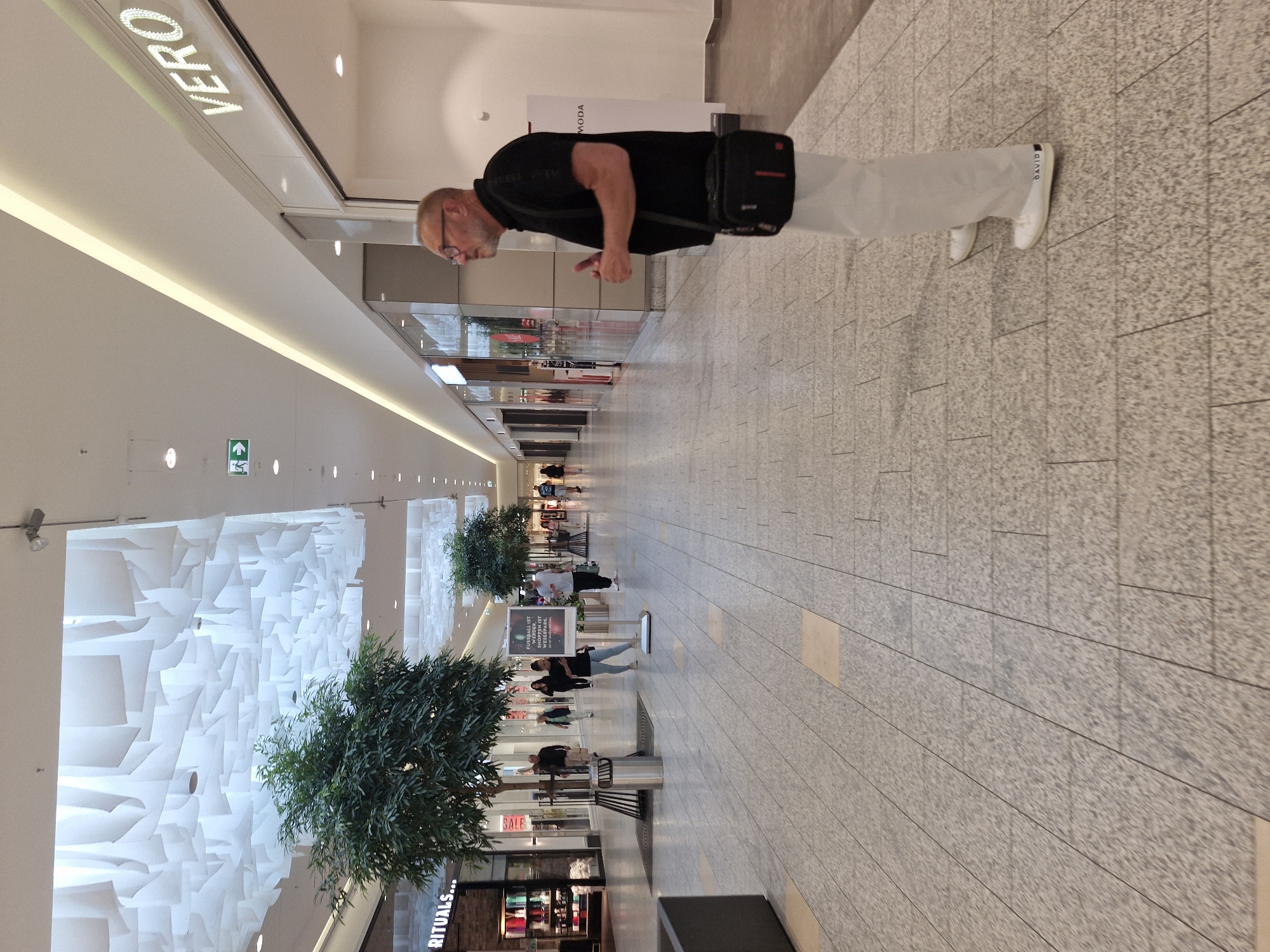 The media file shows a man standing in a modern, well-lit shopping mall. The man is wearing a black t-shirt, light-colored pants, and white sneakers. He has glasses and a short beard. He is wearing a black shoulder bag with a red stripe. His gaze is directed downwards as if looking at something on the floor.
The mall interior features a light-colored tiled floor and a high ceiling with decorative white geometric panels. There are several trees planted in large planters throughout the mall, and shops are visible in the background, some with illuminated signage. The lighting suggests it is daytime, and the indoor setting indicates it's not weather-dependent.
Visible text includes "VERO" prominently displayed on a sign above a shop. Another sign in the distance reads "MODA". Further down the mall, a sign displays "RITUALS..." and another has "SALE". There are also directional signs with arrows and a fire exit symbol. The overall impression is a typical shopping experience in a contemporary European mall.