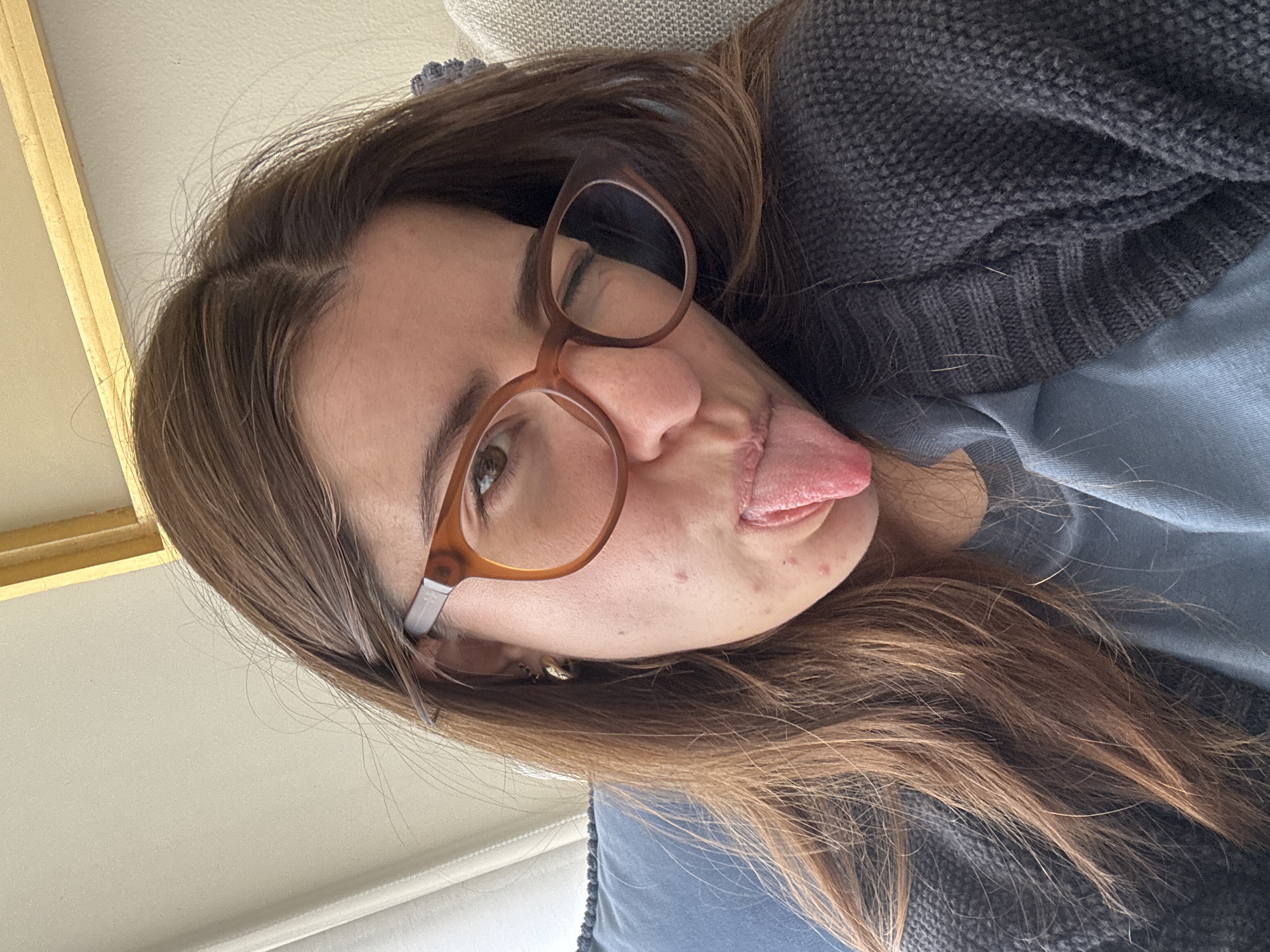 A young woman with brown hair and brown-rimmed glasses is lying down, sticking out her tongue. She is wearing a grey sweater over a blue shirt. The background shows a portion of a wall and furniture, suggesting an indoor location. There is no information in the image to suggest it was taken in Puebla, Mexico.