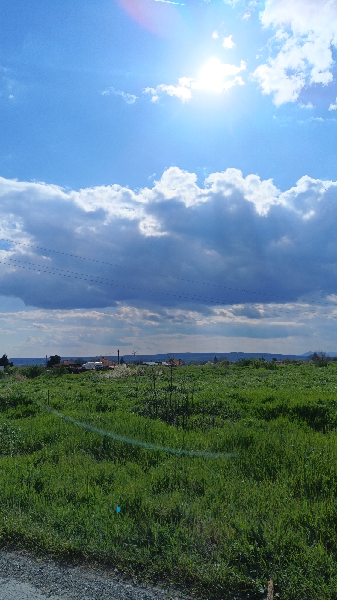The image depicts a rural landscape in Valchin, Bulgaria, under a bright, partially cloudy sky. The sun is visible, positioned high in the frame. A large band of clouds dominates the mid-ground, casting shadows on the land below. Power lines are strung across the sky just above the clouds. In the distance, a cluster of buildings, possibly a small village, is discernible. The foreground is dominated by a field of green grass, with some scrub vegetation scattered throughout. A segment of a gravel road is visible in the extreme foreground.