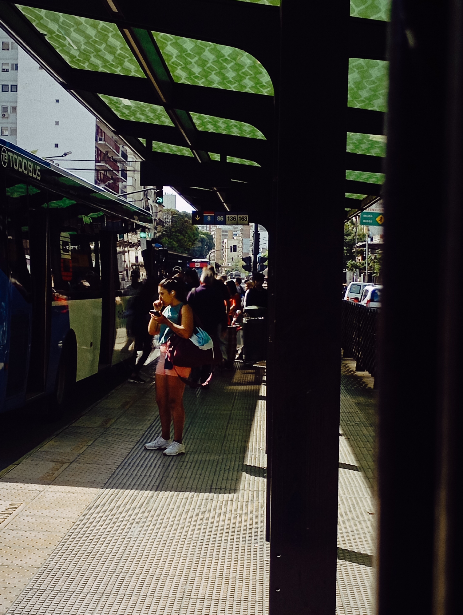 An outdoor scene depicts a bus stop platform in Buenos Aires, Argentina. People are waiting for a bus, the Todo Bus is visible on the left side of the image. A woman wearing a blue shirt and pink shorts is on her phone. The bus stop has a green glass roof. The bus stop platform has rows of patterned blocks. Several buildings can be seen in the background.