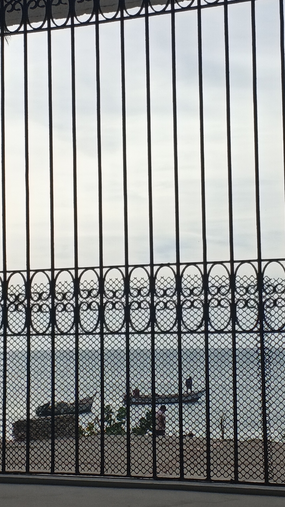 The image presents a view of the bay in Juan Griego, Venezuela, observed through a black metal grille in the immediate foreground. The grille consists of vertical bars, an upper decorative band of interconnected oval shapes, and a lower section with a diamond-patterned mesh.
Beyond the grille, the scene depicts a body of water, characterized by light ripples, extending from the middle ground to the horizon. Above the water, the sky is light grey and appears overcast.
On the water, two small vessels are visible. A darker-colored, indistinct boat is positioned near the left, seemingly close to or partially beached on the shoreline. Further right, a lighter-colored, elongated boat floats on the water. Two human figures are present in this boat; one stands near its right end, while the other is seated or crouched towards the center. On the narrow shoreline between the grille and the water, a third human figure is partially obscured by vegetation, wearing a light-colored shirt and facing the water. Scattered dark objects, potentially rocks or debris, are on the shoreline near the left boat.