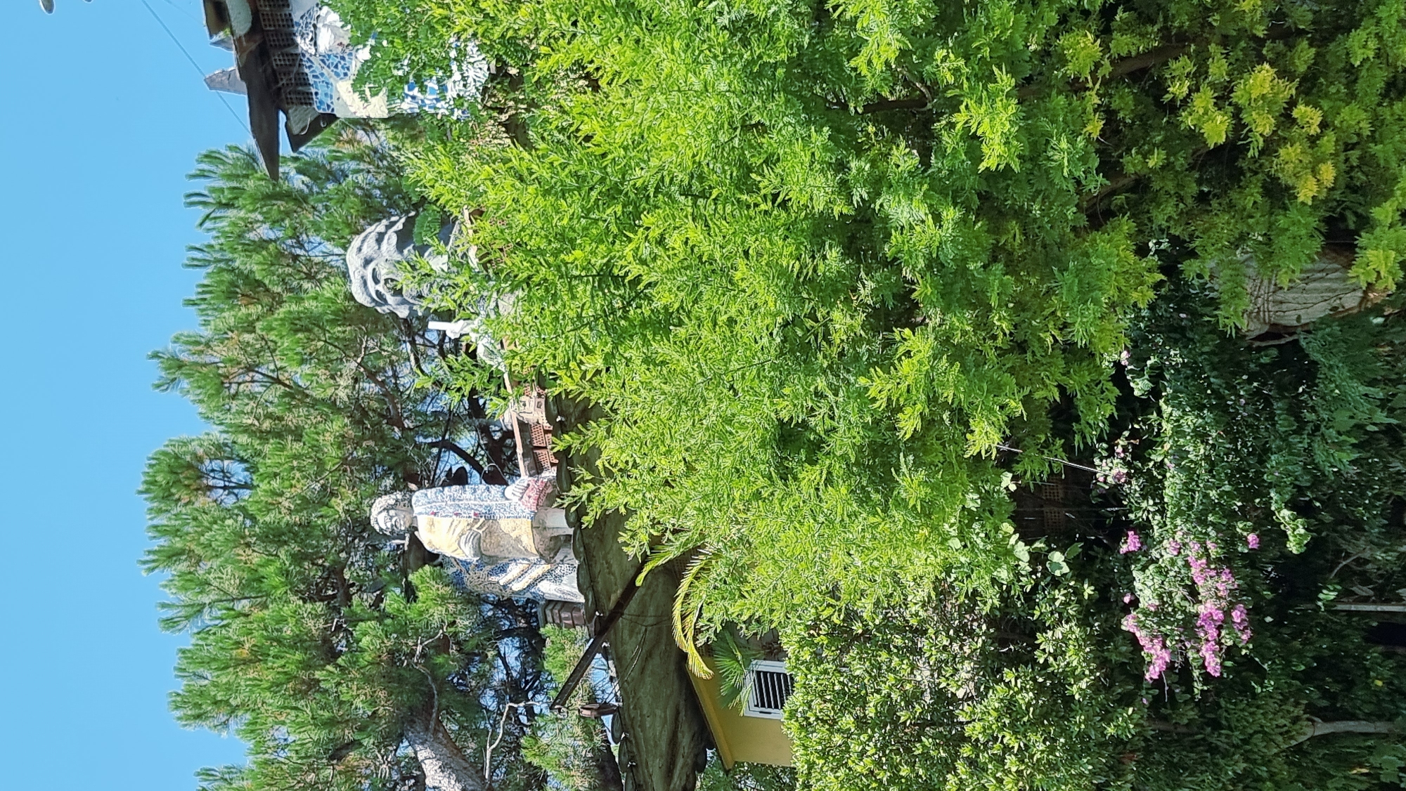 The image depicts an outdoor scene with dense green foliage and trees against a clear blue sky. In the background, partially obscured by branches, are several unusual structures with mosaic tile embellishments and statues. One statue appears to be of a person, while another is a large, sculpted head, possibly of a lion or mythical creature. Below these structures, a portion of a yellow building with a white-shuttered window is visible, also surrounded by greenery. The lower part of the image shows more lush vegetation, including a cluster of small pink flowers. The overall impression is of an eccentric, art-filled property integrated into a natural environment. This scene is located in the city of Sant Joan Les Fonts, Spain.