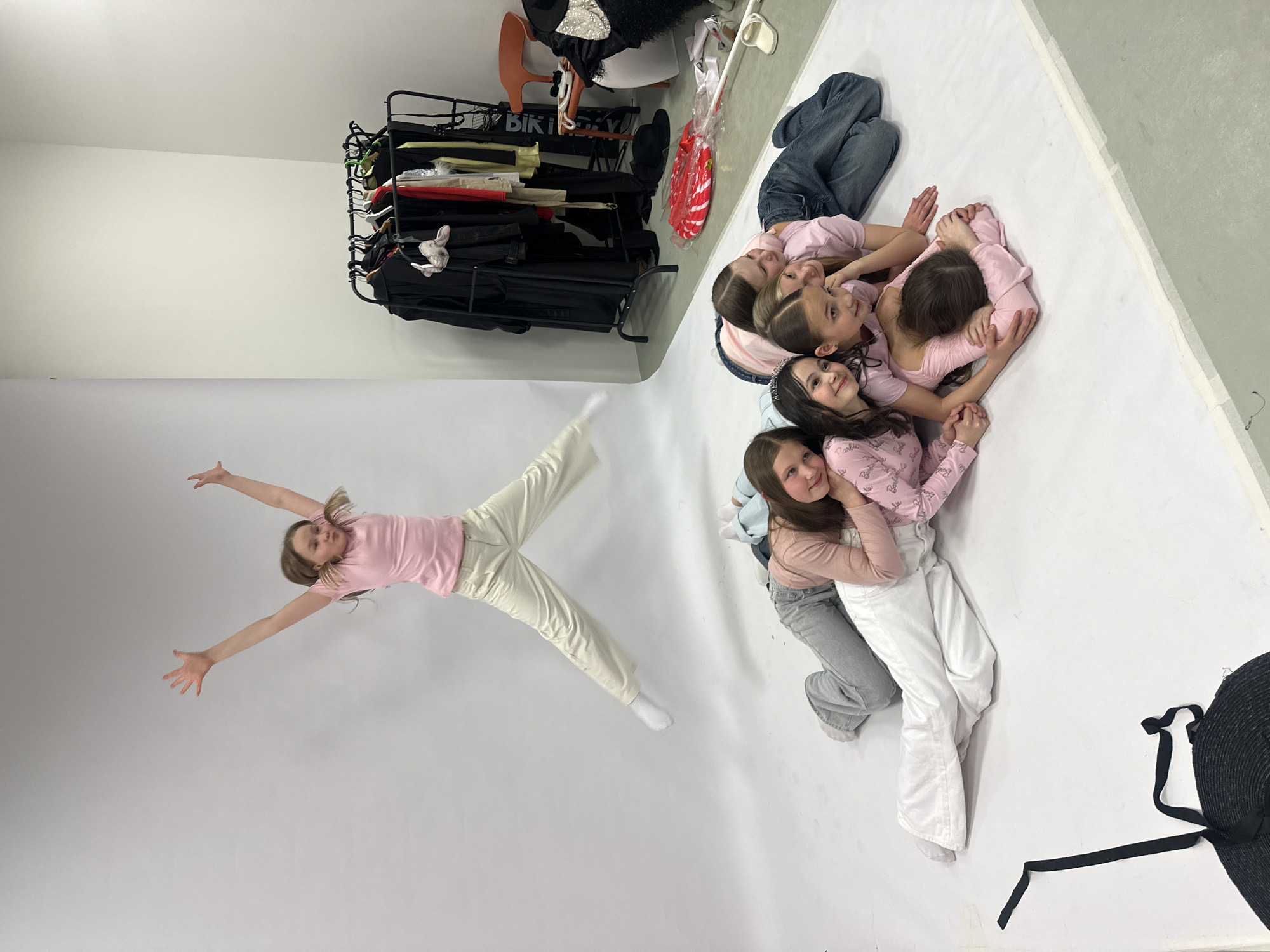 In a studio setting, against a white backdrop, several young girls are arranged in different poses. Six girls are clustered together on the upper right side of the frame, lying in a circular, interconnected formation. They are wearing various shades of pink tops and jeans. To the left of this group, a single girl is positioned with her arms outstretched and legs spread wide, as if in mid-jump or suspended in the air. She is wearing a pink top and white pants.
A black clothing rack filled with garments stands in the upper left corner, with additional items such as an orange chair and a red decoration partially visible behind it. An indeterminate black object is partially visible in the lower right corner.
The scene appears to be a photograph taken in a studio during a photoshoot, with the arrangement of the girls and the presence of clothing racks suggesting a professional or staged environment. The image has been rotated counter-clockwise. There is no information to suggest that the photo was taken in the city of Votkinsk, Russia.