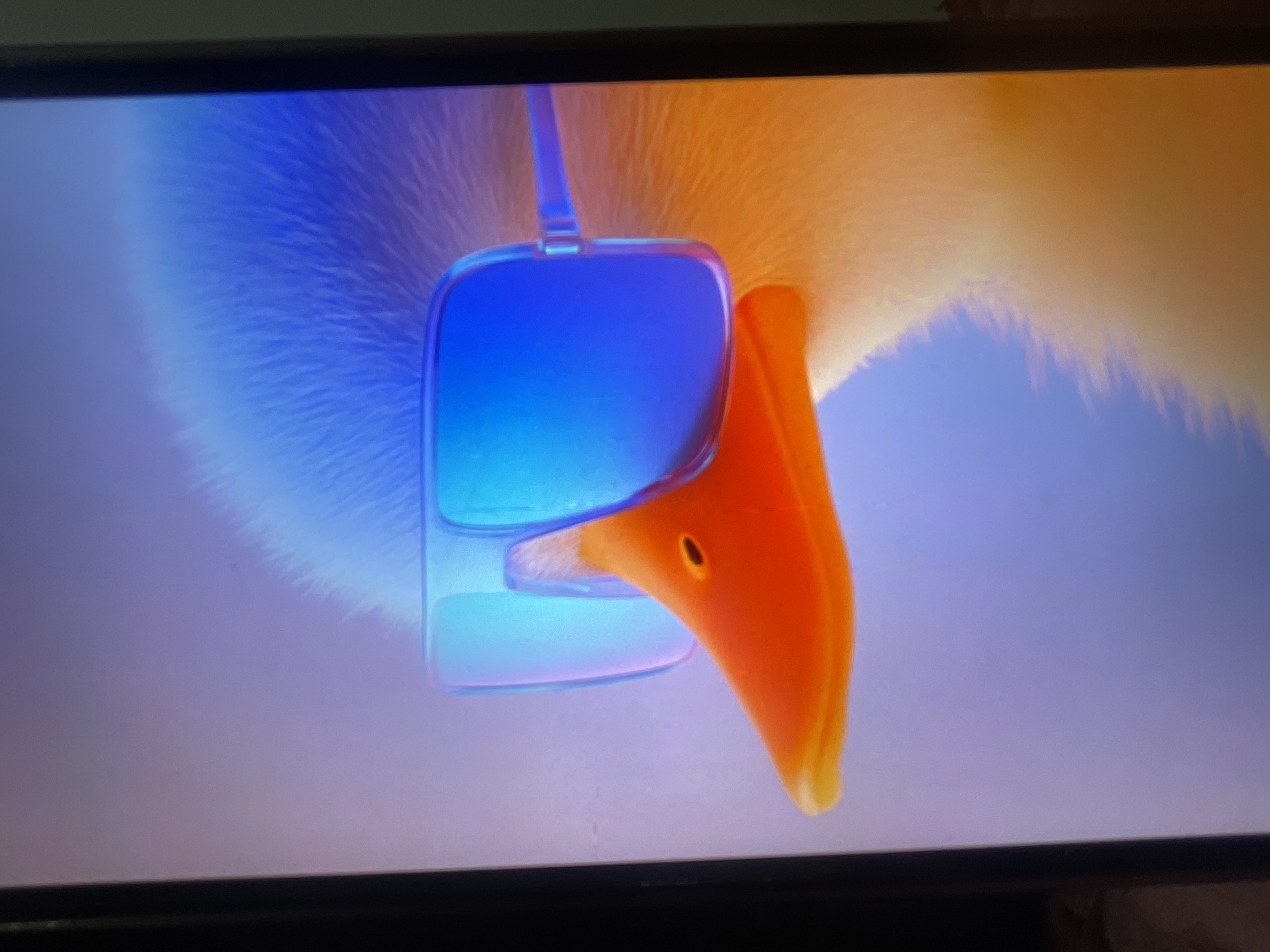 The image shows a close-up of a cartoon duck's face. The duck is wearing large, square, blue sunglasses. Its beak is bright orange and has a small, dark nostril. The duck's head appears to be covered in fluffy blue and yellow feathers. The background is a soft gradient of purple and pink. There is no discernible location in Bishkek City, Kyrgyzstan, visible in this image.