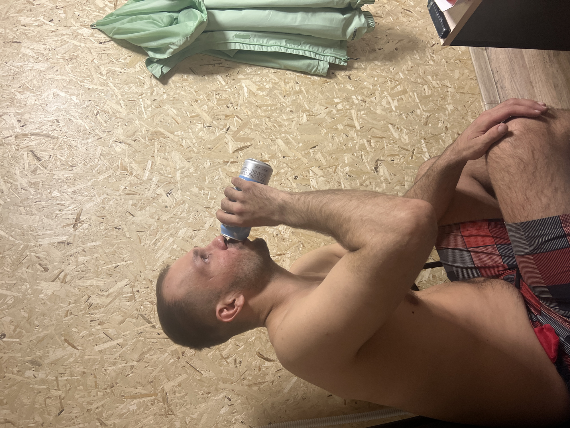 A Caucasian male is drinking from a blue and silver can. He is shirtless and wearing red and black plaid shorts. He is lying on what appears to be a wooden floor. To the left and above the man is a green folded garment. To the right and above the man is a black box and a beige wall. The can has Cyrillic script. Based on the Cyrillic script and the user's request, the location is likely Chaikovsky, Russia.