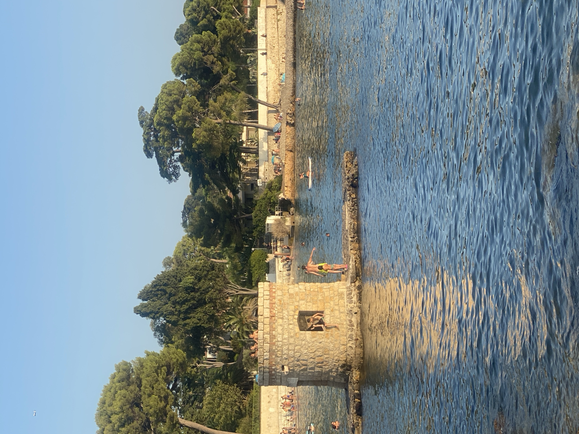 The image captures a sunny day at a beach and water area in Antibes, France, likely near the Fort Carré, given the presence of a stone structure reminiscent of a watchtower. In the foreground, the calm, blue water reflects the sunlight, creating a shimmering effect. A small, rocky island with a stone tower stands in the middle ground. Several people are visible interacting with the water and the tower. On the tower, a person in a yellow swimsuit is striking a pose, while another person in dark swimwear sits in the tower's window opening. A man and a young girl in a swimsuit are standing on the rocks adjacent to the tower. Further out in the water, individuals are swimming and one person is paddleboarding. The background reveals a sandy beach lined with people relaxing under umbrellas and trees. A wall separates the beach from a vegetated area with numerous tall trees, typical of the Mediterranean flora. In the upper left corner, a distant airplane is visible against the clear blue sky. The overall scene depicts a vibrant and leisurely atmosphere at a coastal location.