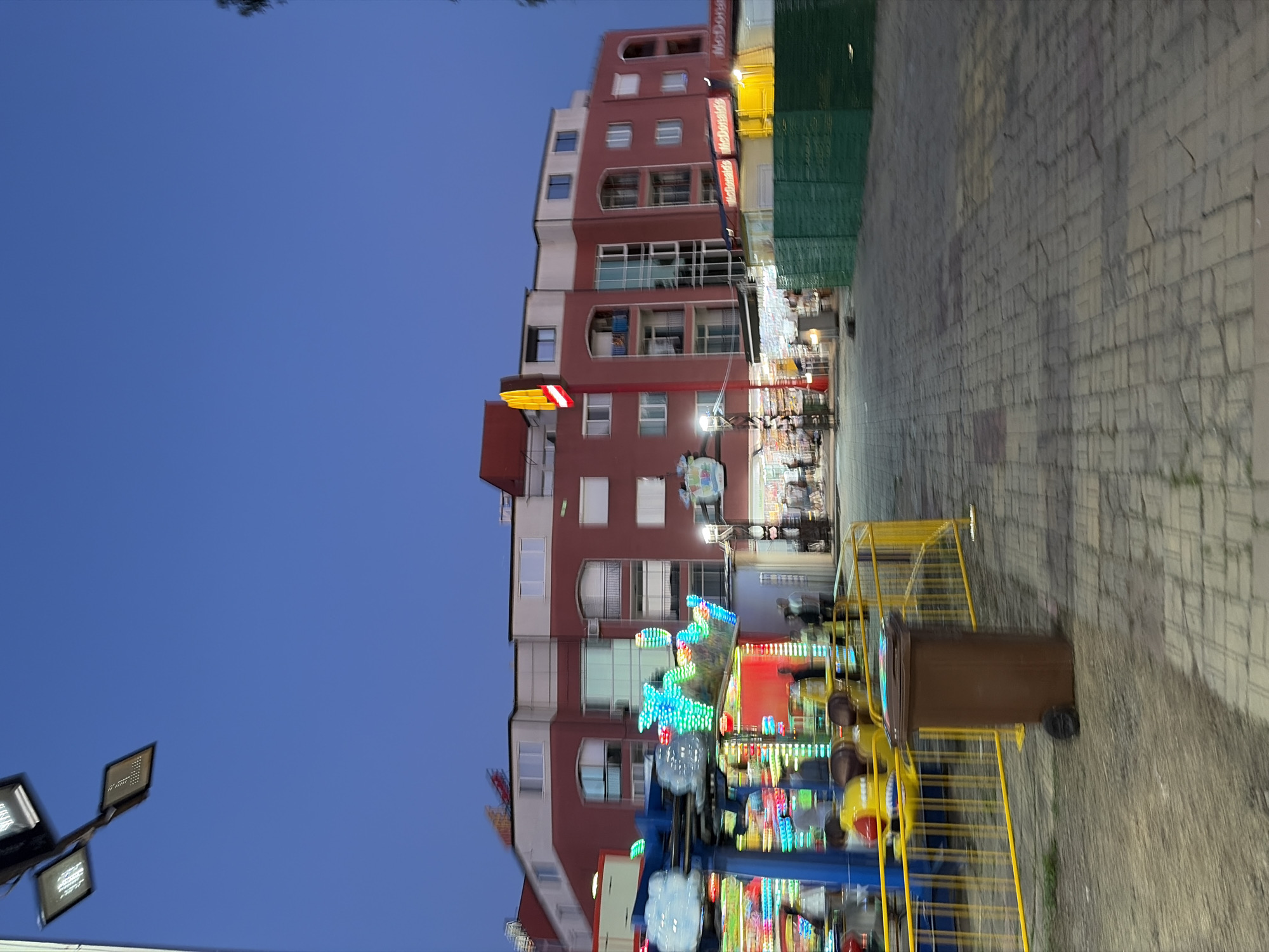The image shows an outdoor scene at dusk, possibly in a public area like a boardwalk or amusement park, with a building in the background and a ride in the foreground. The sky is a deep blue, transitioning from daylight to evening.
In the foreground, a brightly lit amusement ride with flashing colorful lights is visible. Yellow metal barriers section off the ride area. A brown trash can stands to the right of the ride. The ground is paved with cobblestone-like bricks.
The building in the background is a multi-story structure with a red brick facade and white sections. It appears to have balconies and windows. Several signs are visible on the building, including a prominent McDonald's logo.
A few individuals are present in the scene, mostly blurred due to motion or low light. They appear to be walking or standing near the building and the ride. The overall atmosphere suggests a lively, public gathering place.