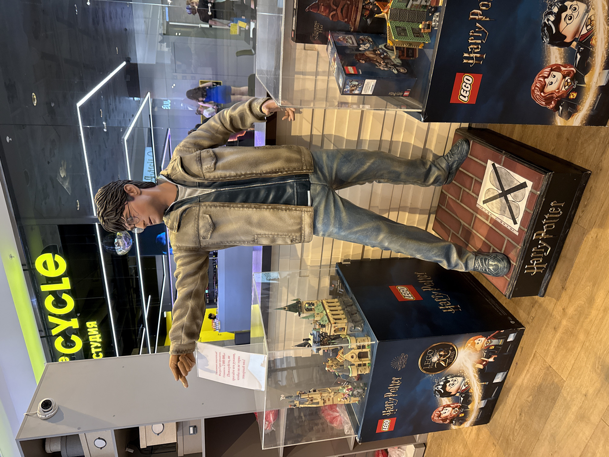 The image shows a life-sized Harry Potter figure displayed in what appears to be a retail or exhibition space. The figure, wearing a jacket and jeans, is positioned above several Lego Harry Potter sets stacked on boxes. A sign is placed in front of the statue in Russian. In the background, there is a store called "CYCLE" in Moscow, Russia, with additional retail spaces visible. The setting suggests a commercial environment.