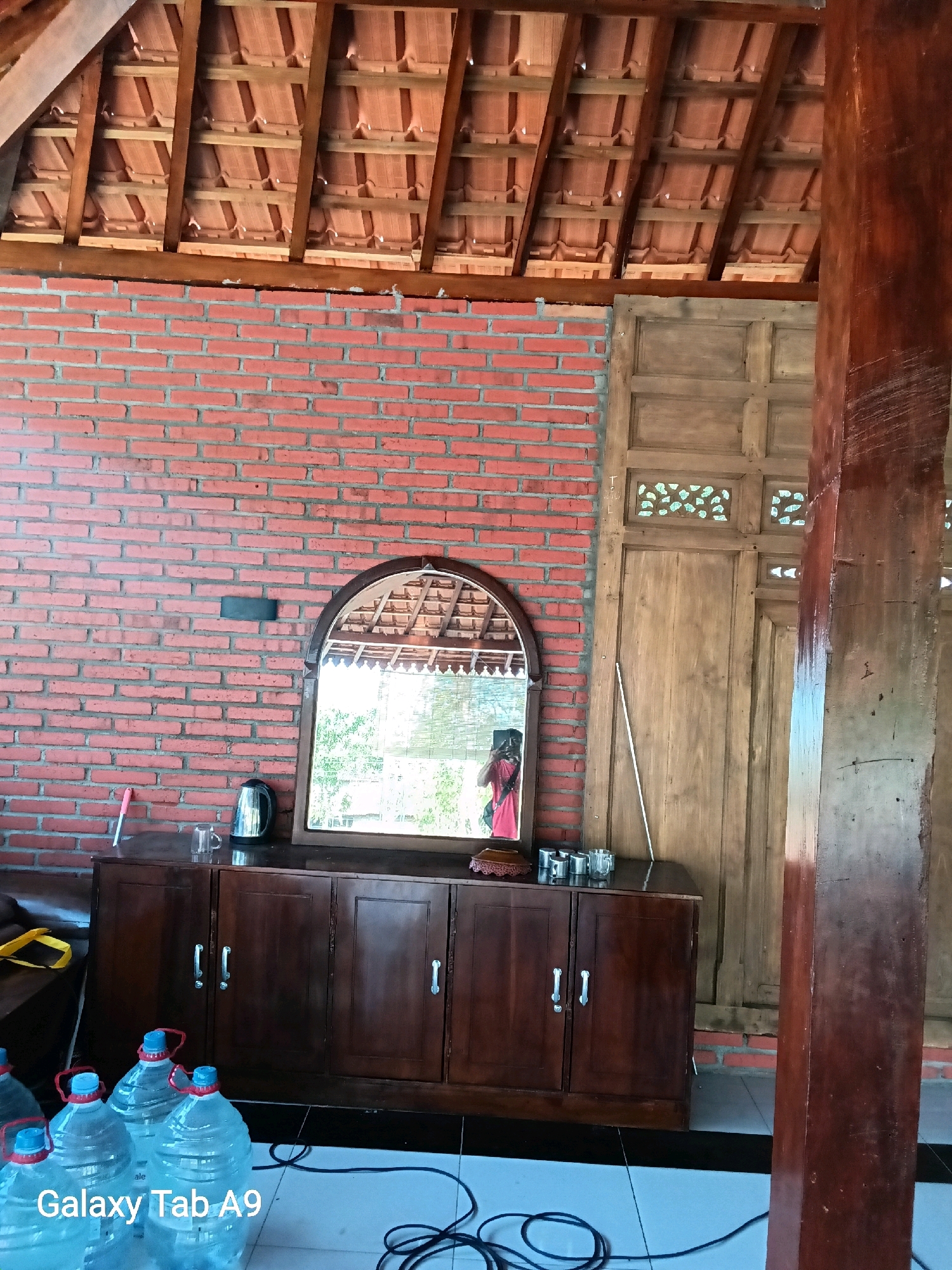 The image shows an interior scene of a building with a rustic aesthetic, possibly a restaurant or guesthouse in Trenggalek, Indonesia. The walls are constructed from red brick, and the ceiling features exposed wooden beams supporting terracotta roof tiles.
Dominating the left side of the image is a dark wooden cabinet with a mirrored top. The mirror reflects the interior, including a person holding a phone and taking the photo, and the view of the outdoors through a window. On the cabinet's surface, a black electric kettle, a glass, and a pink object, possibly a cleaning brush, are visible.
To the right of the cabinet, there is a large wooden door with intricate carvings in the upper panels. A long, thin white stick leans against the doorframe. A thick, reddish-brown wooden pillar stands prominently on the right edge of the frame, indicating structural support.
In the foreground, several large plastic water jugs with blue caps are arranged on a tiled floor. Black electrical cords are visible on the floor, one of which is connected to the water jugs. A partial view of a yellow object, possibly a bag or cushion, is visible on the far left. The text "Galaxy Tab A9" is superimposed on the lower left portion of the image.