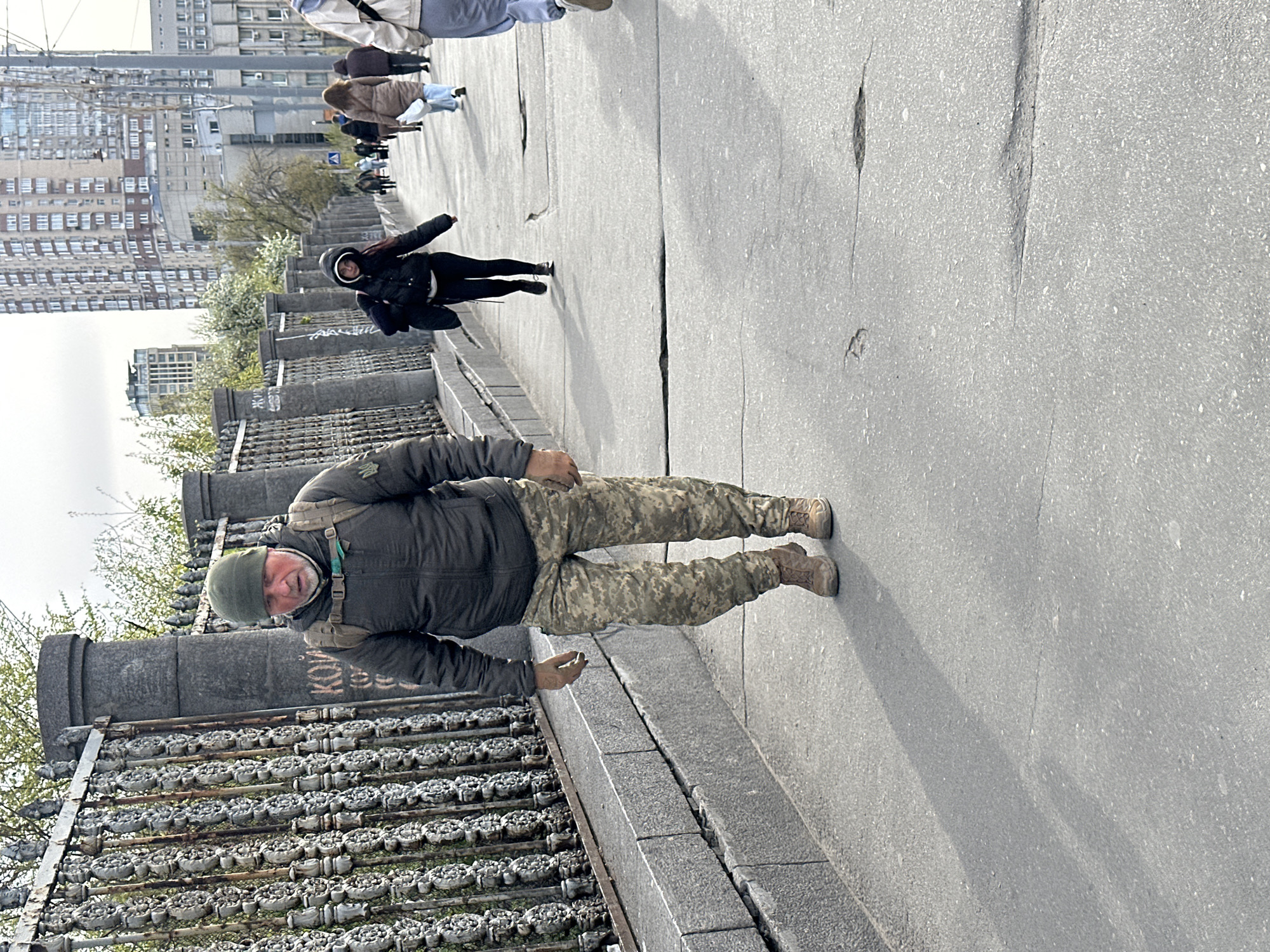 The image captures an urban outdoor scene, likely an elevated walkway or bridge, in Kyiv, Ukraine, during the daytime. The sky appears overcast or partly cloudy, casting soft shadows.
In the foreground, a man is the primary subject, dressed in military-style clothing. He wears a dark green padded jacket, camouflage trousers, and brown combat boots. A green beanie hat covers his head, and he has a short beard. A tactical backpack is visible on his shoulders. He holds a small, light-colored object, possibly a stone or a nut, in his right hand. He is looking directly towards the viewer with a neutral to slightly serious expression.
To the man's left, a decorative, dark grey fence or railing runs along the edge of the walkway. It features repetitive circular or floral patterns, possibly made of concrete or metal. Behind this railing are large, cylindrical concrete structures, one of which has faint, painted letters, possibly "KOB" or "KOBM," visible as graffiti. Further beyond, green trees, some with white blossoms, indicate springtime.
The man stands on a wide, light grey paved surface that shows several cracks and imperfections. Further down this path, other people are visible, walking away from the camera. One woman in a black jacket and black pants is more distinctly seen. In the background, numerous multi-story apartment buildings, typical of Soviet-era architecture, line the skyline, confirming an urban setting. The overall impression is one of everyday life in a city, possibly with undertones of the ongoing conflict due to the man's attire.