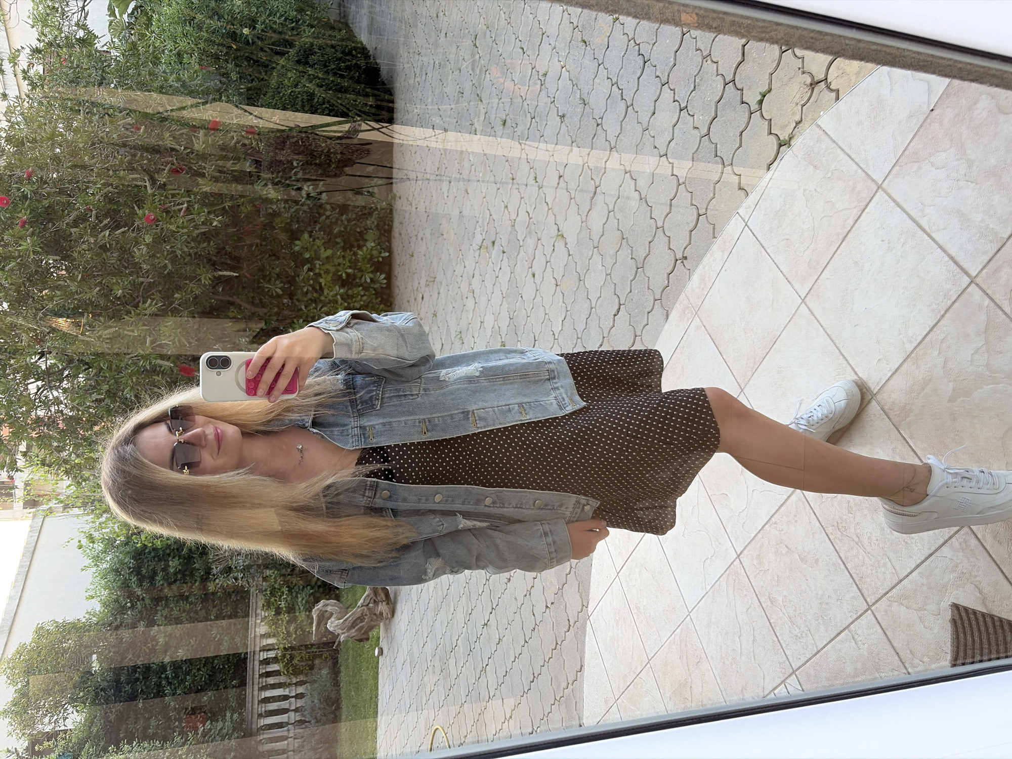 A fair-skinned female with blonde hair and sunglasses is taking a selfie in a mirror. She is wearing a brown dress with white polka dots and a distressed denim jacket. She is also wearing white sneakers. The reflection in the mirror shows an outdoor setting, with foliage and light-colored pavers. A building is seen in the background of the reflection. The location is Budva, Montenegro.