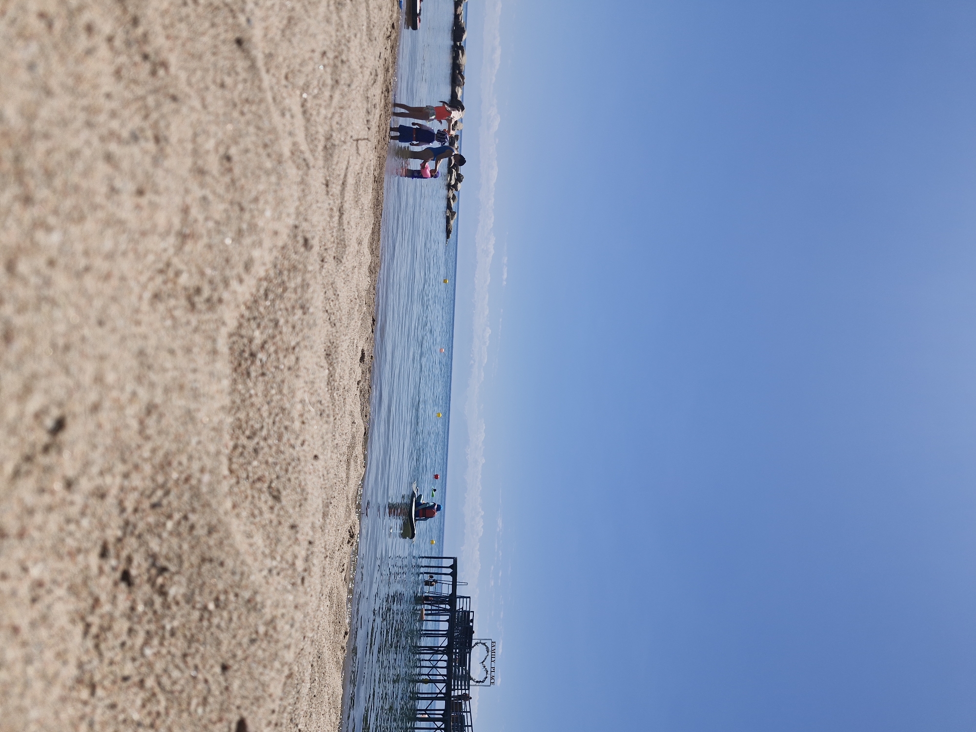 The image shows a serene beach scene at Lake Issyk-Kul in Bulan-Segettu, Kyrgyzstan. The foreground is dominated by a close-up view of the sandy beach, with the grains of sand in sharp focus. Beyond the sand, the clear blue water of the lake stretches to the horizon, meeting a vast, cloudless blue sky. In the mid-ground, a group of people are visible enjoying the water. Three children and an adult stand near the shore, with two children partially submerged in the shallow water. Another individual is seen on a paddleboard further out in the lake, wearing a life vest. In the background, a wooden pier extends into the lake, adorned with a heart-shaped floral decoration and a sign reading "FAMILY PLACE." The pier is lined with people, and several yellow buoys are scattered in the water. Rocky structures are visible along the shoreline to the left of the frame. The overall atmosphere is one of peaceful recreation and summer enjoyment.