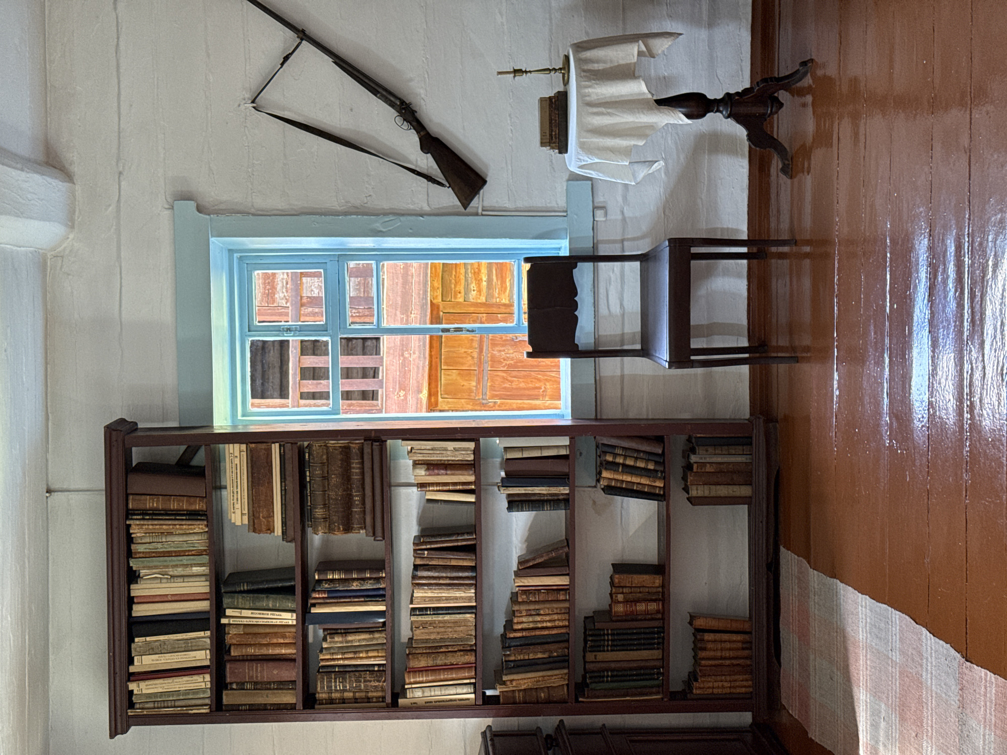 The image depicts an interior scene, likely a room in a historical building in Shushenskoye, Russia. A tall, dark wooden bookshelf filled with numerous old books dominates the left side of the frame. To the right of the bookshelf is a light blue-framed window revealing an exterior view of wooden structures. A dark wooden chair is positioned in front of the window, and next to it, a small, dark wooden table draped with a white tablecloth holds a stack of books and a brass candlestick. A rifle is mounted diagonally on the white-painted wall above the table. The floor is made of polished wooden planks, reflecting the light. A portion of a striped rug is visible on the left side of the floor. The room has white, rough-hewn walls and a white-painted ceiling with visible beams. There are no people present in the image.