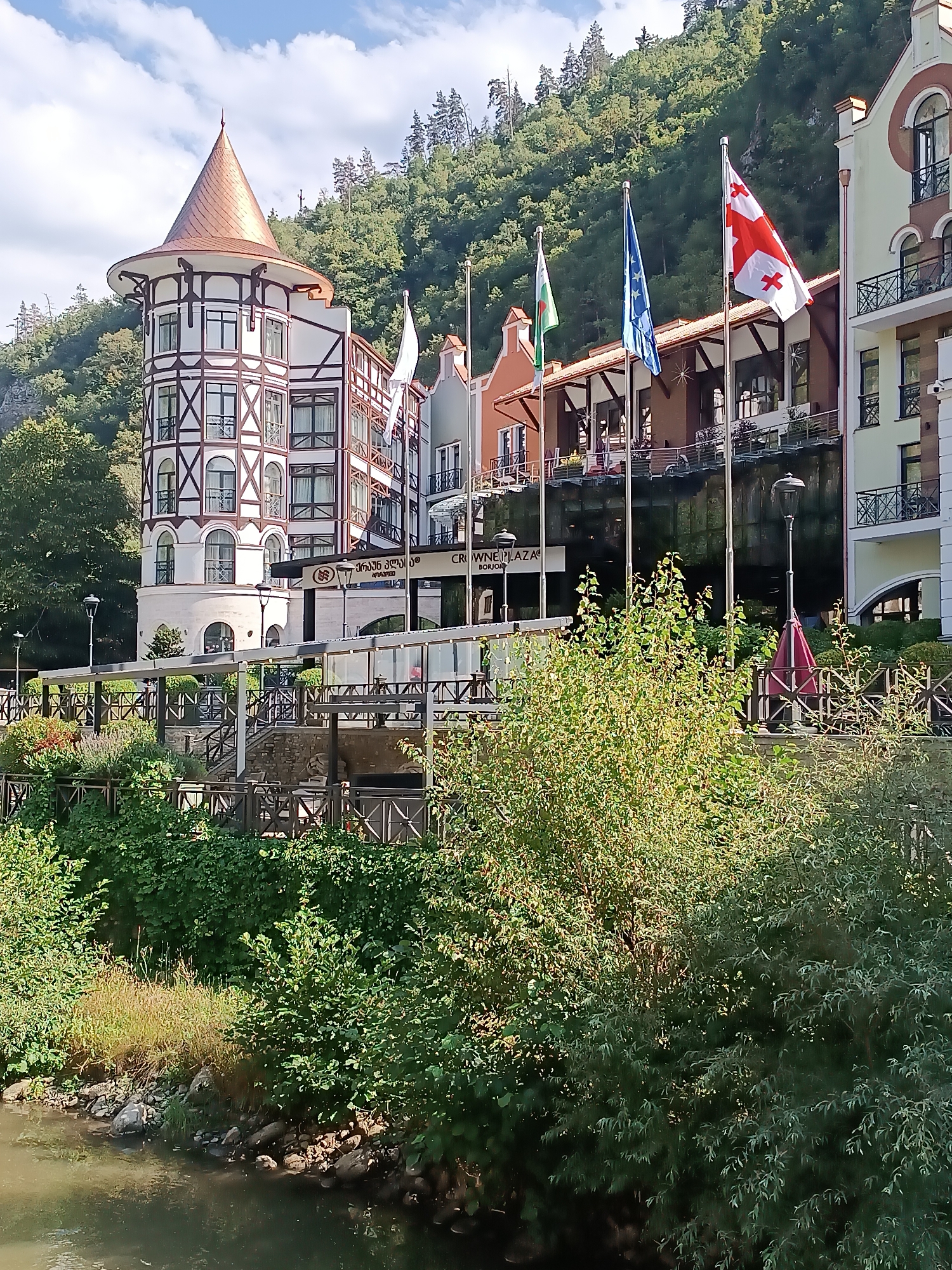 The image depicts a view of the Crowne Plaza Borjomi hotel in Borjomi, Georgia, situated amidst a lush, green hillside and bordering a body of water in the foreground. The hotel features a distinctive European architectural style with a prominent circular tower topped by a copper-colored conical roof. The facade of the hotel is a mix of white and dark timber framing, with numerous windows and balconies. Several flags are visible, including the flag of Georgia, the European Union flag, and what appears to be a regional or organizational flag.
The foreground of the image shows a calm body of water, likely a river, with rocks and vegetation along its bank. A wooden walkway or deck with railings extends along the middle ground, with steps leading up to the hotel. Dense foliage and trees cover the hillside behind the hotel, with a clear sky and scattered clouds above. The scene is bathed in sunlight, suggesting it was taken during daytime. No people are clearly visible in the image, but the overall impression is one of a well-maintained resort or hotel in a natural setting.