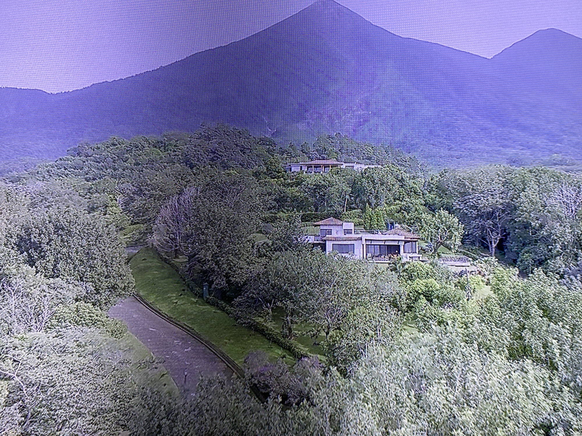 This media file depicts a scenic view of a large, modern house nestled amidst lush greenery on a hillside. In the background, a majestic, purple-hued mountain range dominates the skyline, suggesting a mountainous or volcanic landscape. The house itself appears to be a multi-story structure with a red-tiled roof, large windows, and balconies, indicating a luxurious residence. A paved road curves through the landscape, leading towards the house. The scene is bathed in soft, natural light, possibly indicating a clear day with the sun at a moderate angle, suggesting either early morning or late afternoon. The overall atmosphere is serene and tranquil, emphasizing the harmonious integration of the man-made structure with the natural environment. There are no visible people or any text in the image.