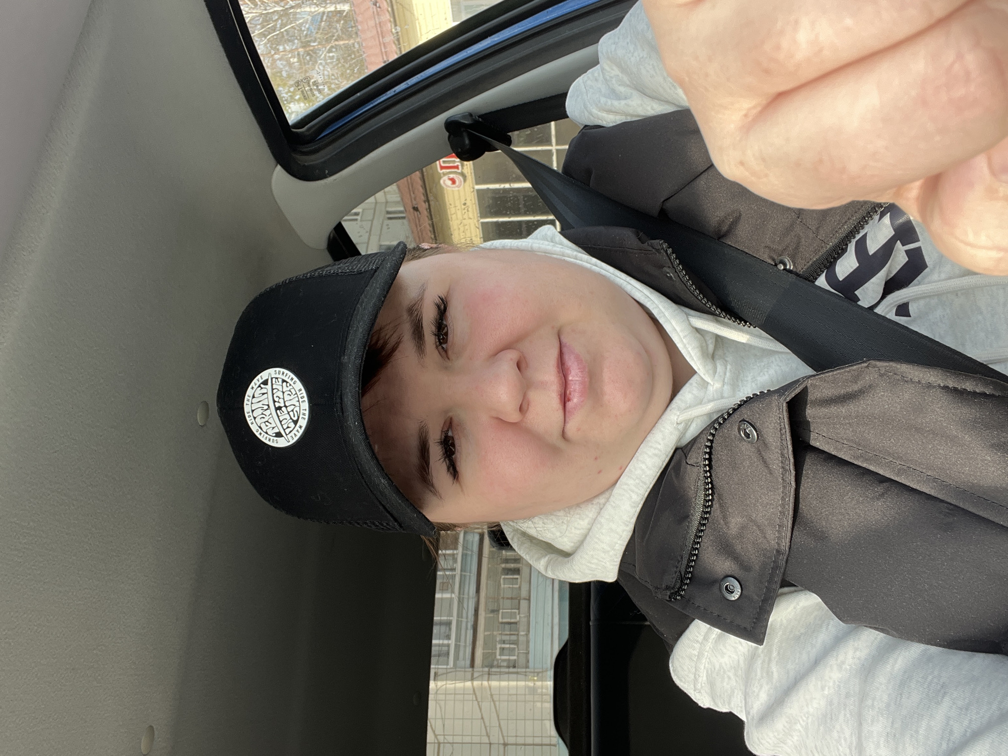 A medium close-up, eye-level selfie depicts an adult individual, appearing female, seated in the driver's seat of a vehicle in Chaikovsky, Russia. The person, with fair complexion and dark hair mostly covered, is wearing a black baseball cap with a white circular logo featuring "RIDE THE WAVE SURFING" around the perimeter and "PERMIA WE LOVE SURF" centrally. They are dressed in a light grey hooded sweatshirt under a black zippered sleeveless vest, secured by a black vehicle seatbelt diagonally across the chest and shoulder. The individual has visible eyeliner and mascara, looking directly at the camera with a slight smile. The right hand, partially clenched, is visible in the lower right foreground.
Through the vehicle's windows, the urban environment of Chaikovsky is visible. To the left, a multi-story, light-colored (possibly white or beige) building with numerous windows and enclosed balconies typical of residential or institutional architecture is discernible. To the right, through the passenger-side window, another building with a yellow facade, a reddish roof, and a red sign with partially visible white Cyrillic characters, including "Опт" (indicating "Wholesale"), is seen. Bare deciduous trees are present in the background, suggesting late autumn, winter, or early spring. The vehicle's interior includes a light grey headliner and a blue door trim on the right.