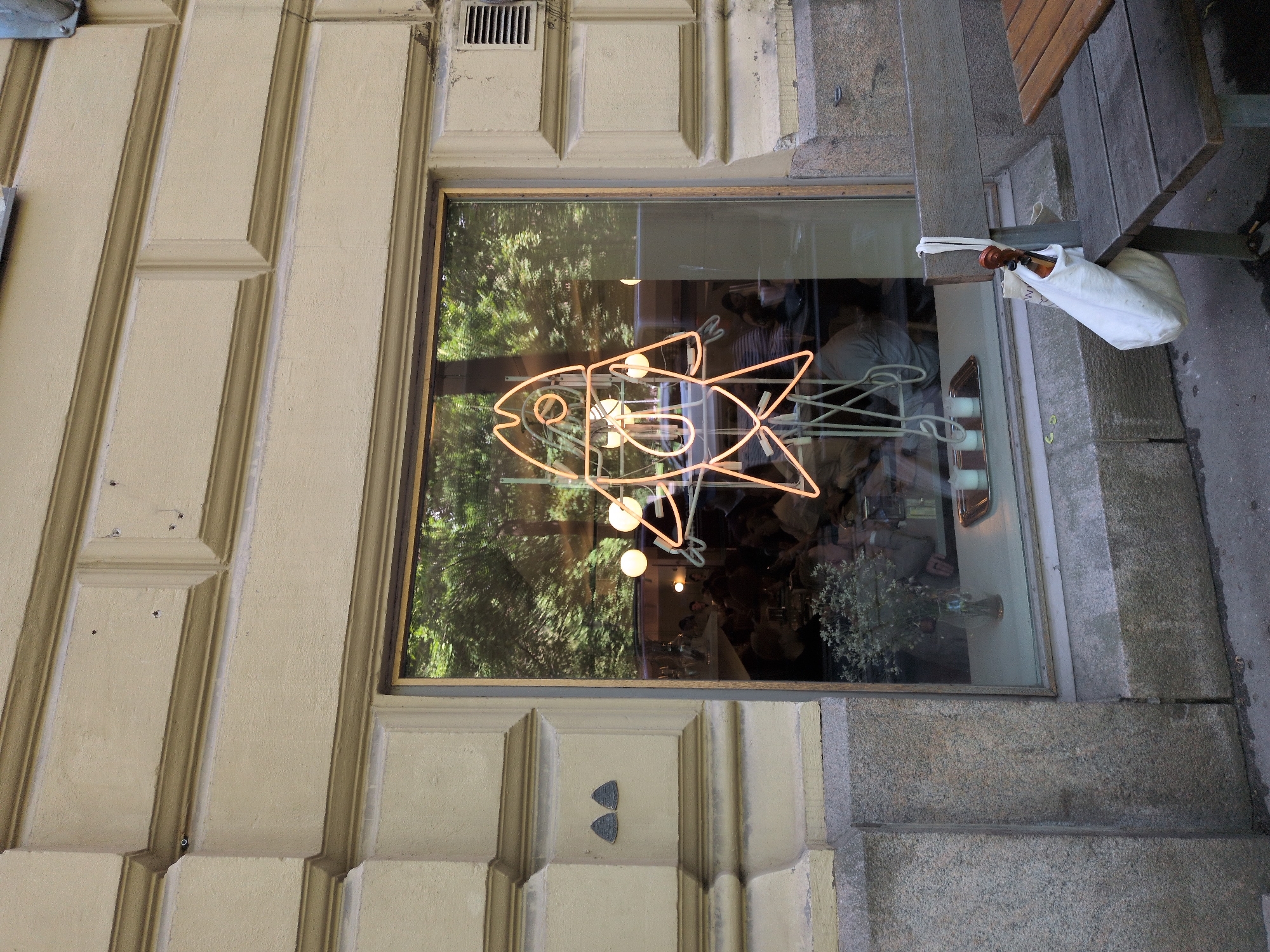 A neon fish sign is illuminated inside a window of a building with beige, textured walls and decorative stone accents. The window reflects trees and interior seating areas with patrons. Outside the window, a wooden bench with a white canvas bag and a violin case hanging from it is visible. Part of a wooden table is also in view. The location is the street-level exterior of a building in Helsinki, Finland.