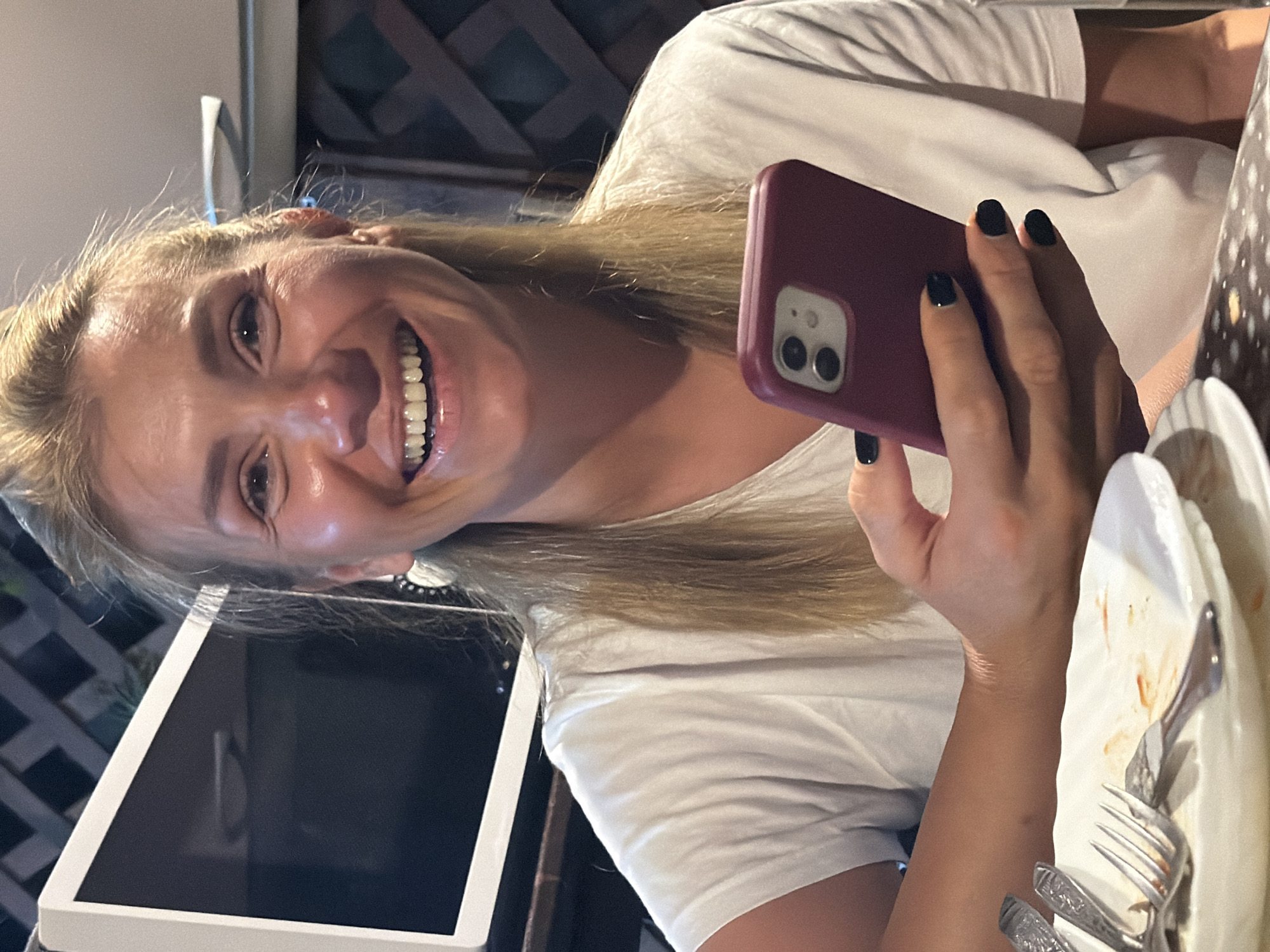In Sochi, Russia, a woman with blonde hair, wearing a white t-shirt, smiles broadly as she holds a burgundy smartphone in her right hand. Her nails are painted black. In the foreground, a white plate with remnants of food and silverware is visible. The background features a dark lattice-like structure and a white electronic device, possibly a monitor.